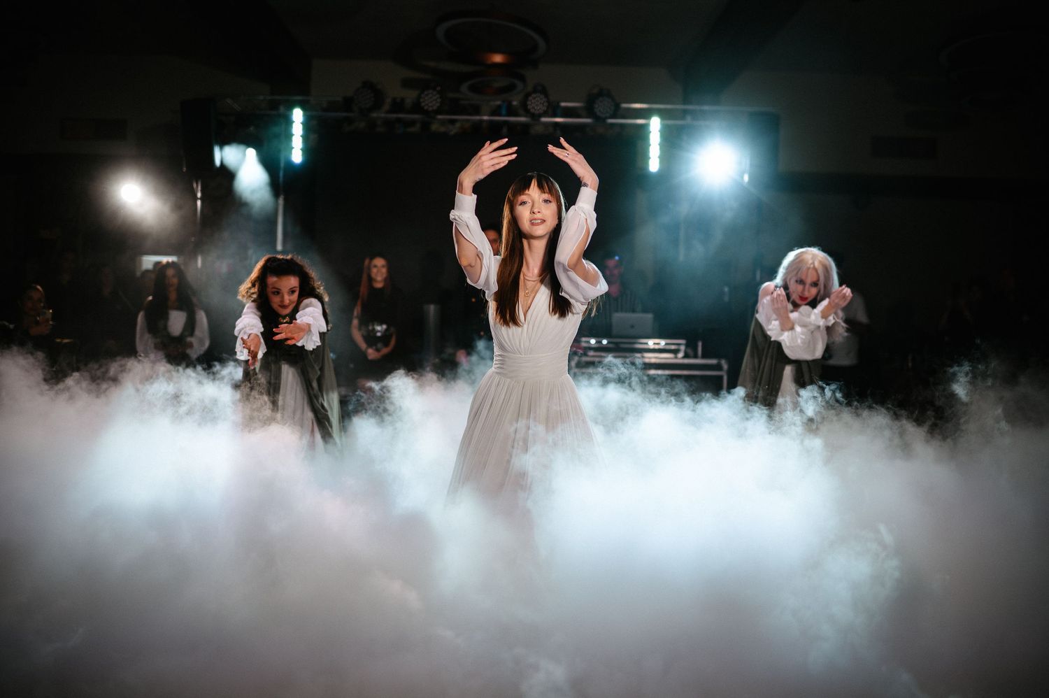 Bride in white dress performs choreographed dance with bridesmaids surrounded by billowing fog on stage.