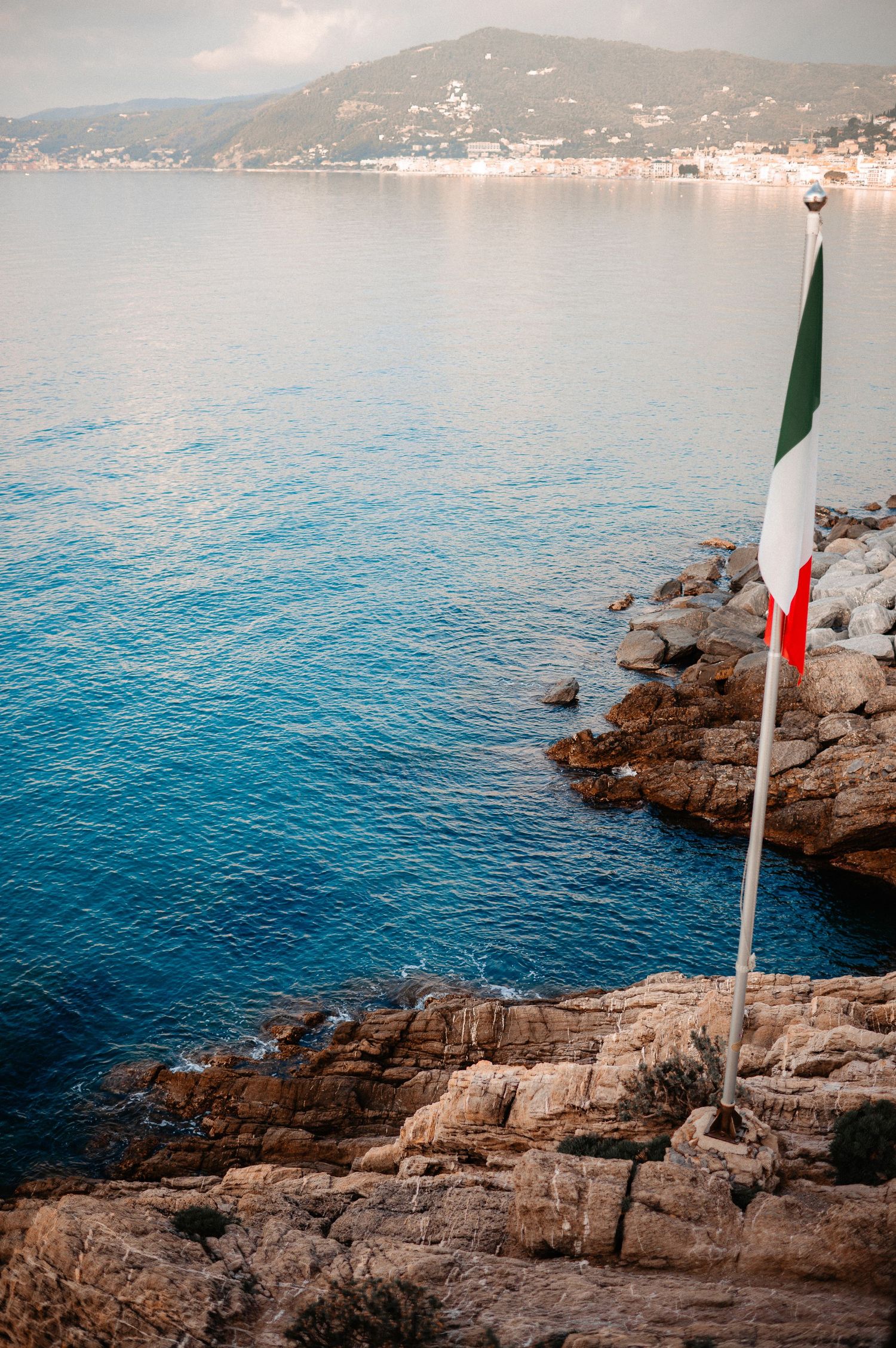 Italian flag on rocky Mediterranean coastline with blue sea and hillside town in background.