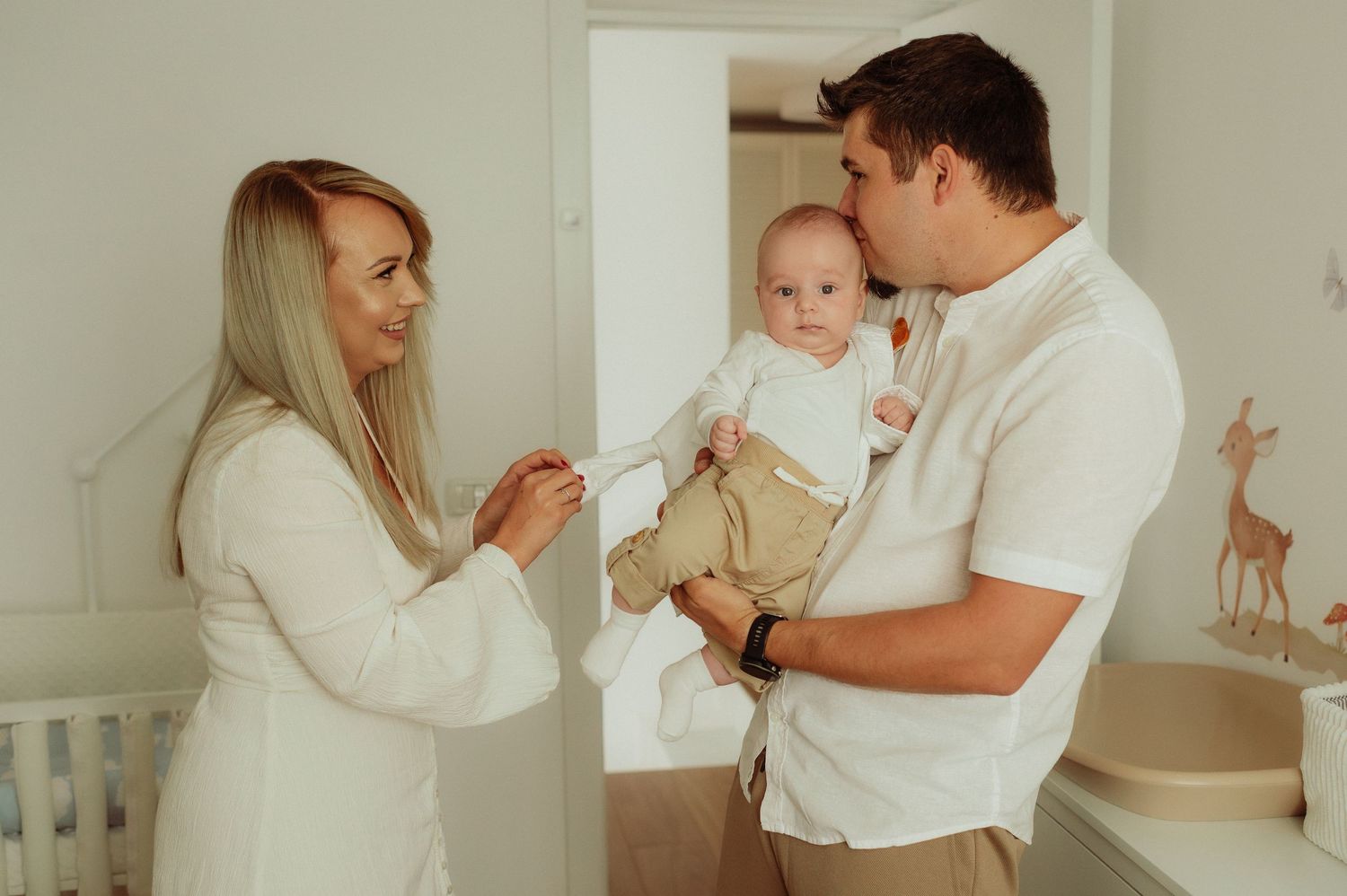Young parents with blonde hair holding newborn baby together in white nursery room.