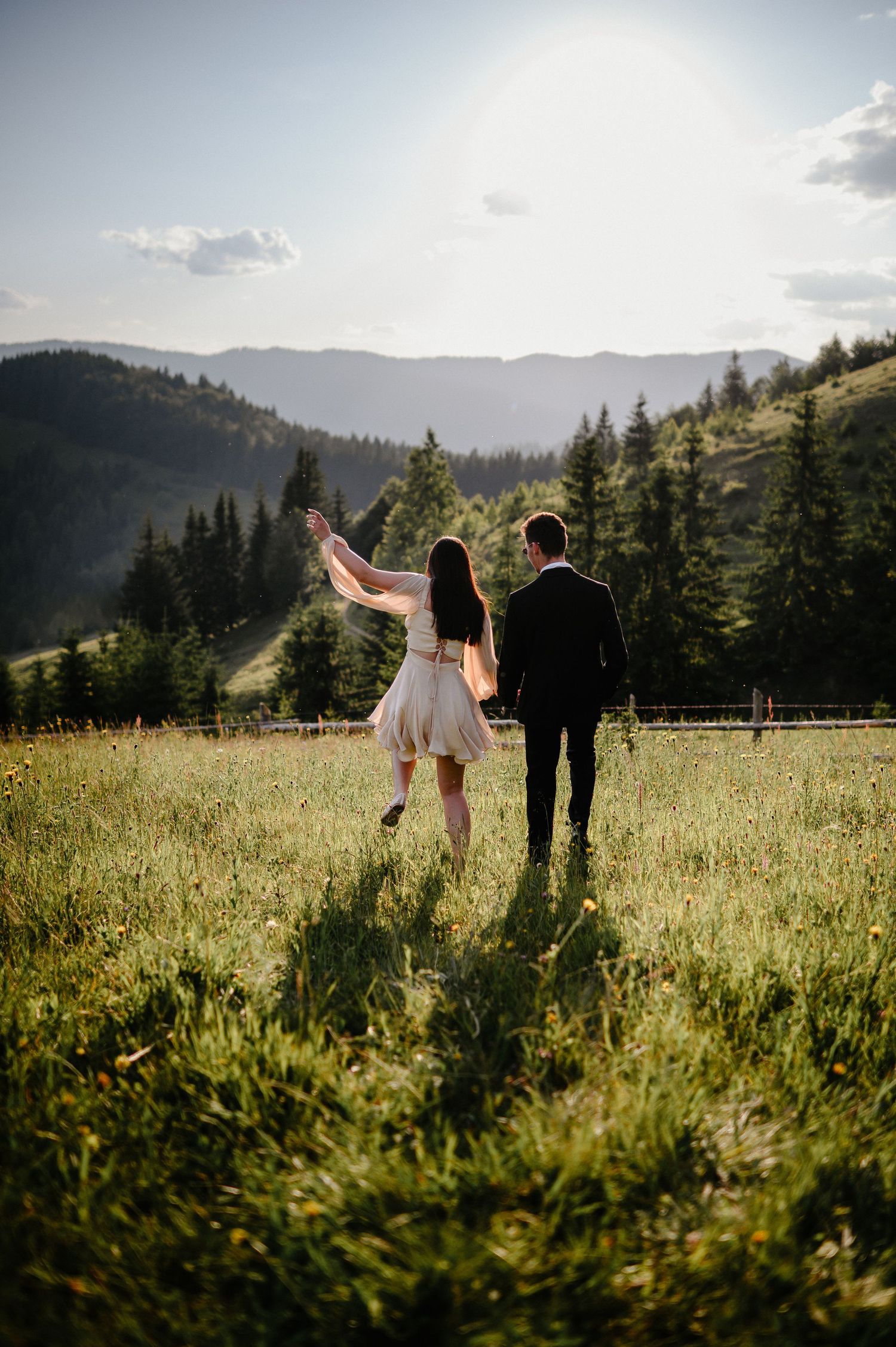 Couple walking hand in hand through a sunny mountain meadow, woman in cream dress raises her arm.