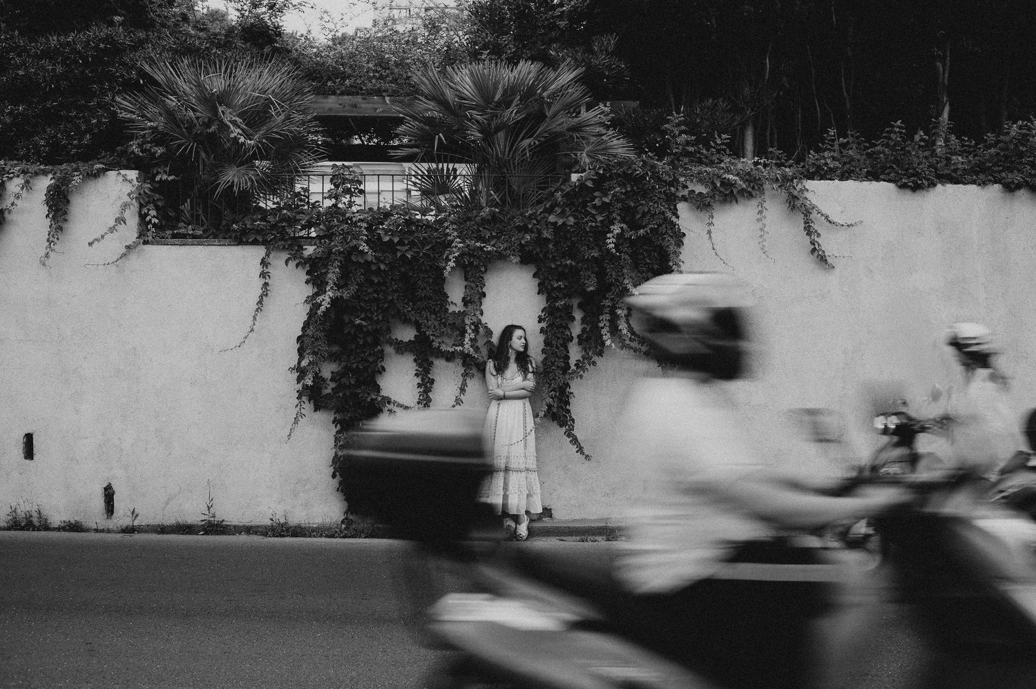 Black and white photo of a woman in white dress standing by an ivy-covered wall with blurred motorcycles.