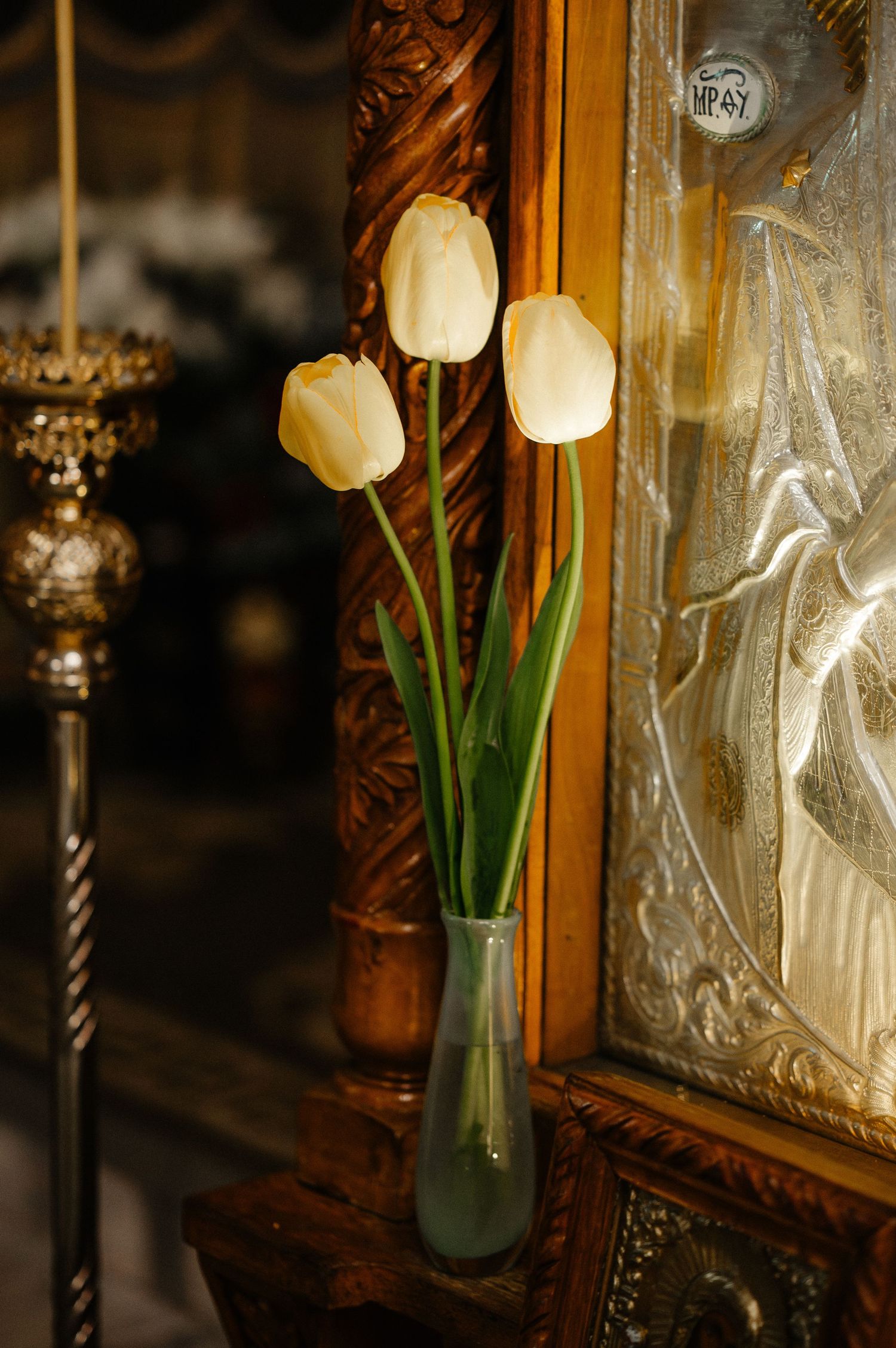 Three white tulips in a glass vase beside an ornate golden Orthodox icon frame.
