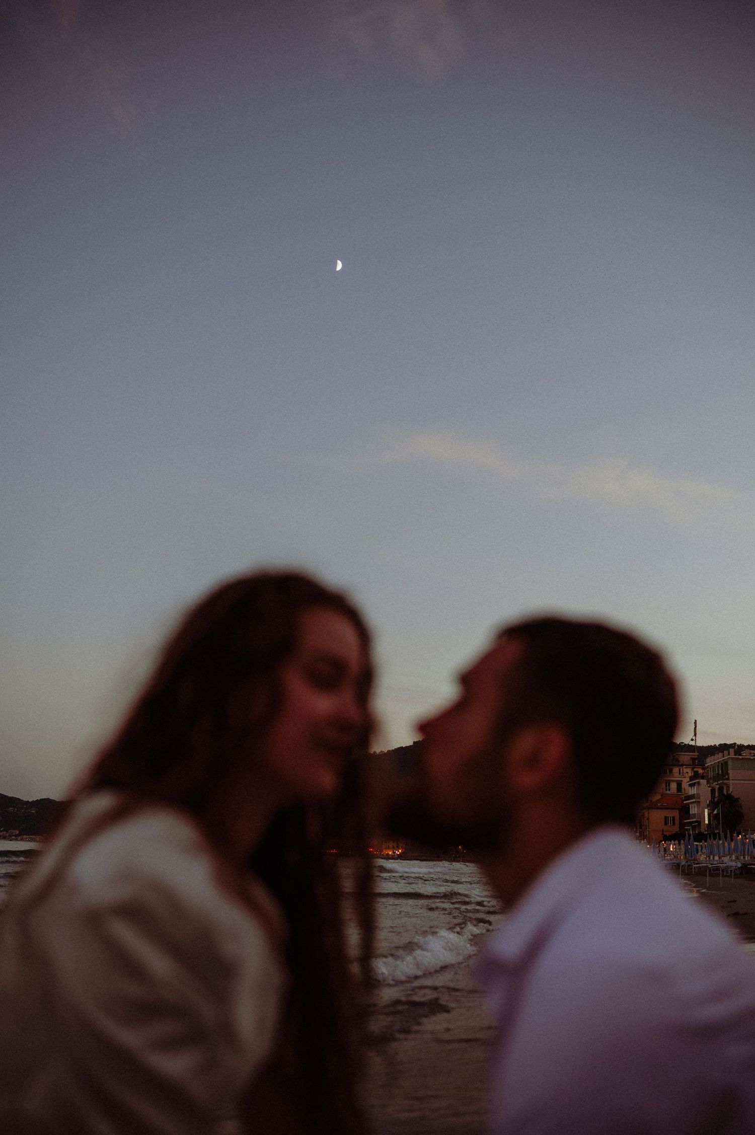 Couple sharing a kiss on beach at twilight with crescent moon visible in dusky blue sky above.