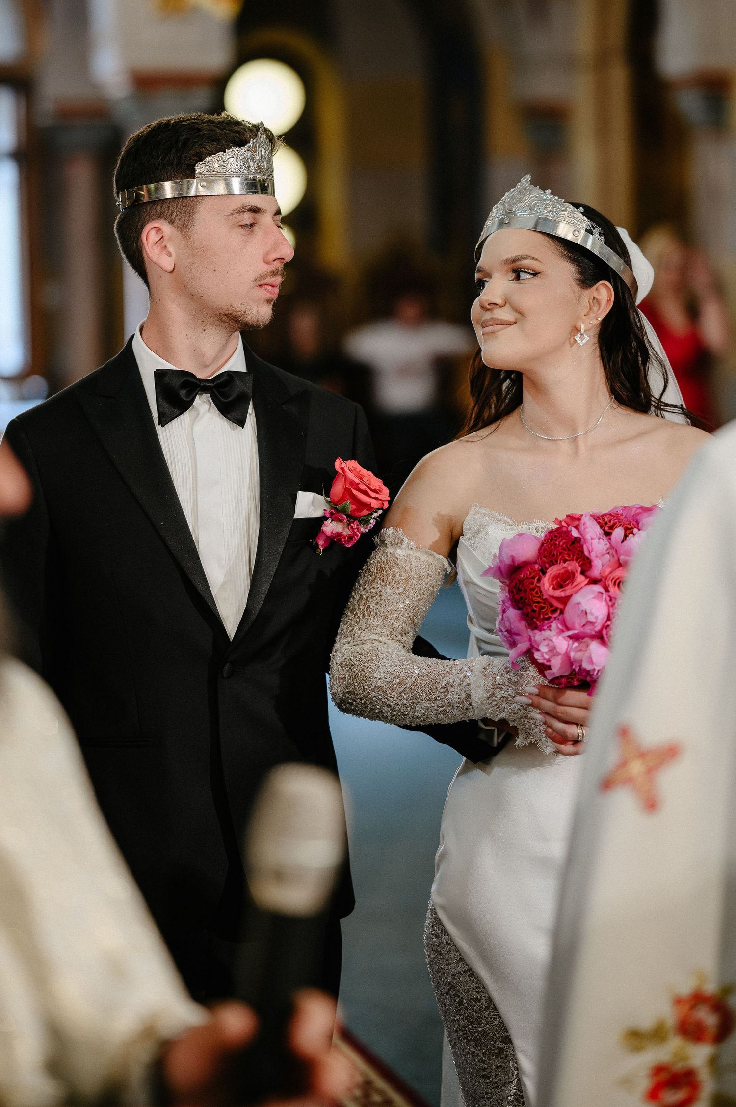 Bride and groom wearing silver Orthodox crowns exchange glances during church wedding ceremony.