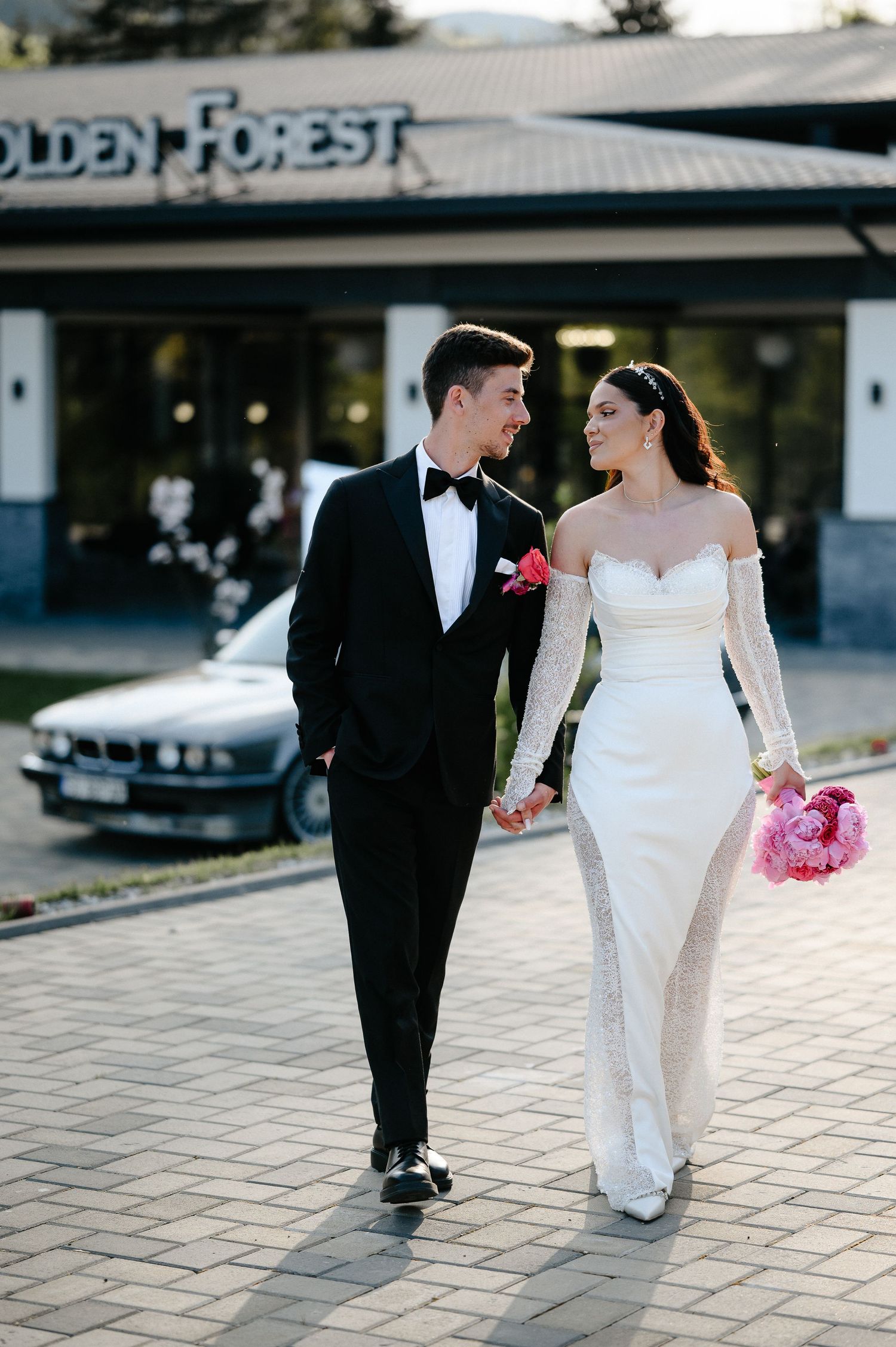 Bride and groom walking hand in hand outside Golden Forest venue, bride in white off-shoulder gown holding pink bouquet.