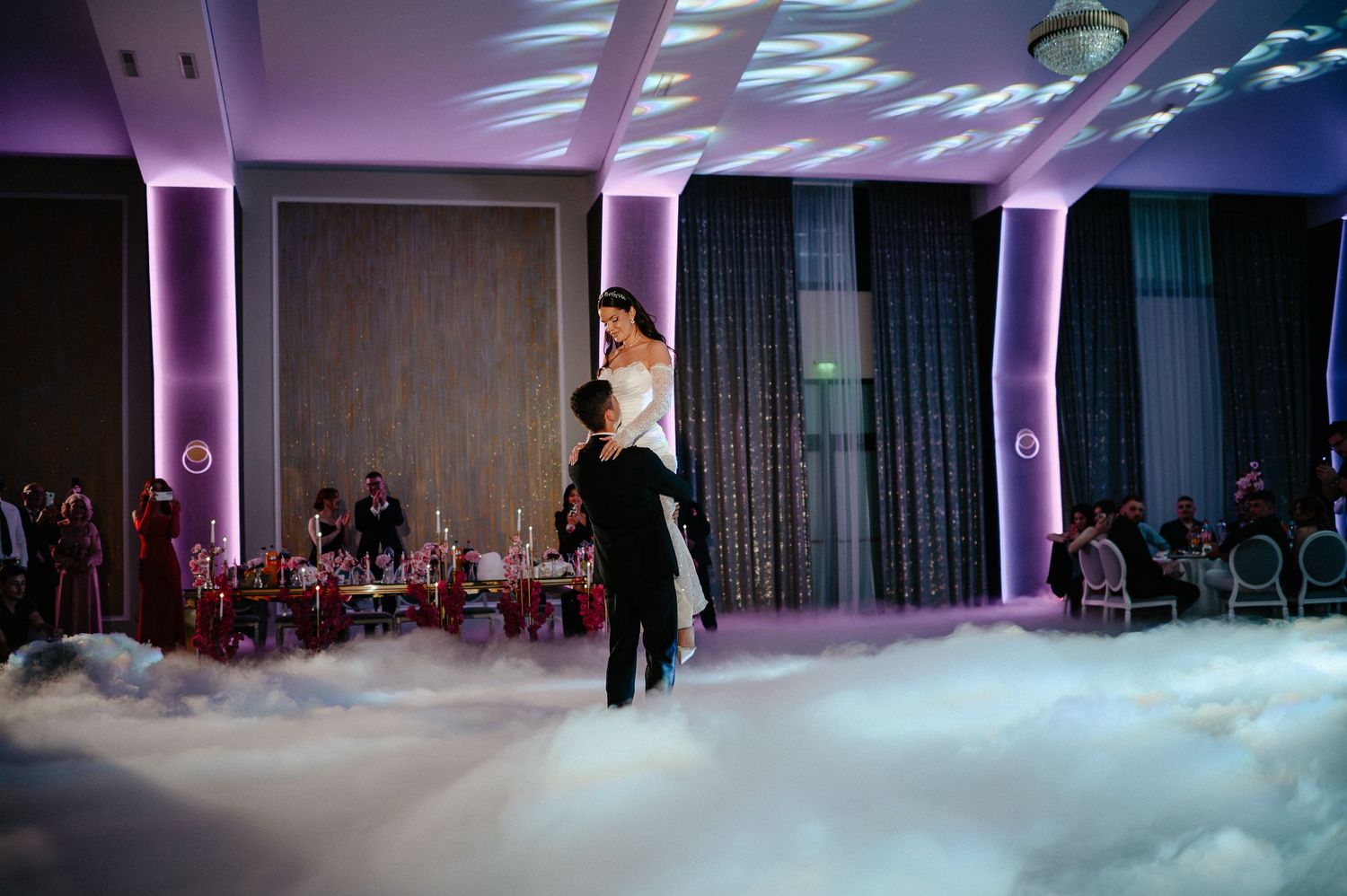 Groom lifts bride high during spectacular first dance on cloud-covered dance floor at elegant wedding.