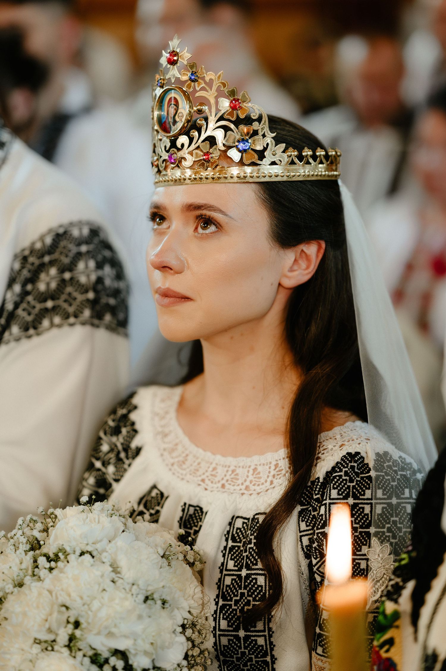 A bride wearing an ornate gold crown and traditional embroidered dress holds white flowers at ceremony.