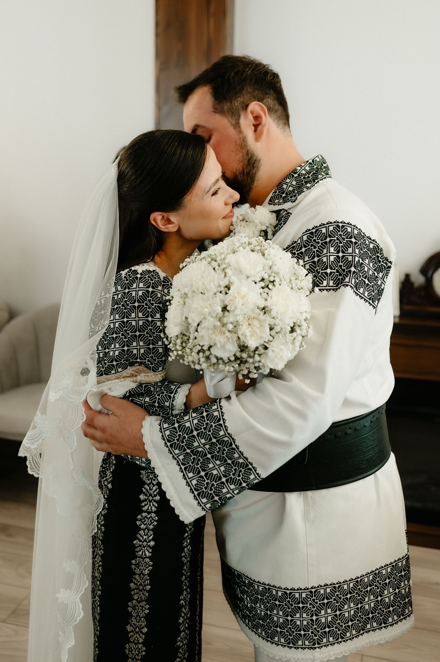 A couple in traditional embroidered black and white attire embrace holding white flowers at wedding.