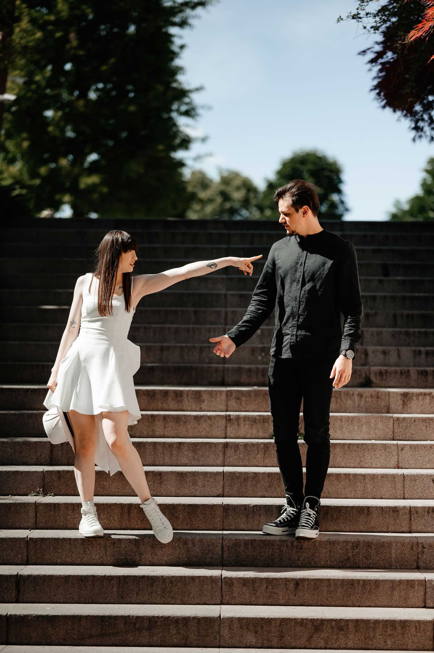 Playful couple on stone stairs, woman in white dress reaching out to touch mans chin, both smiling.