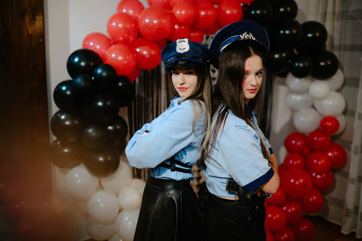 Two women dressed as police officers pose back-to-back before red, black, and white balloons.