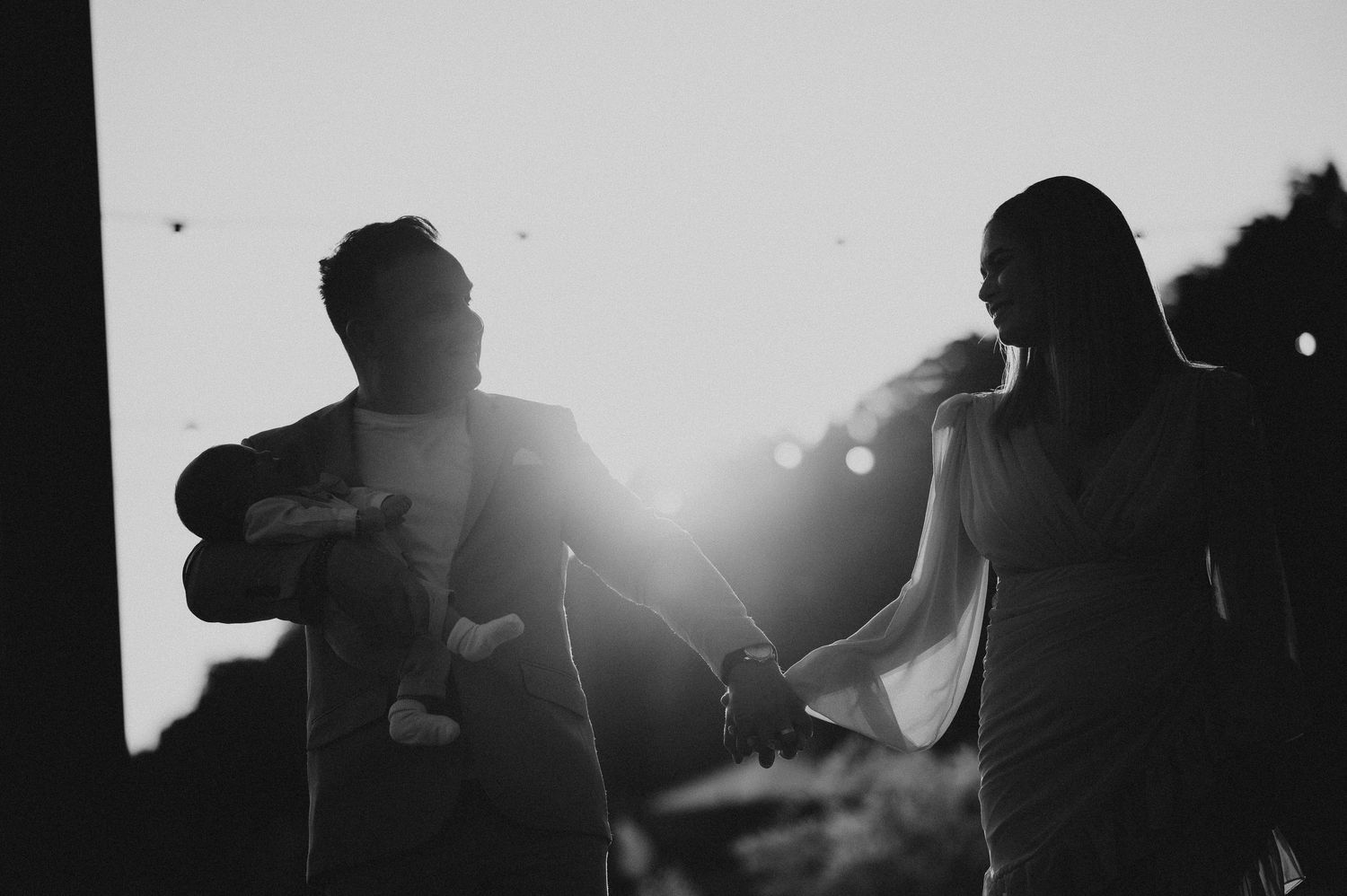 Black and white silhouette of a couple holding hands with a baby against a bright backlit sunset.