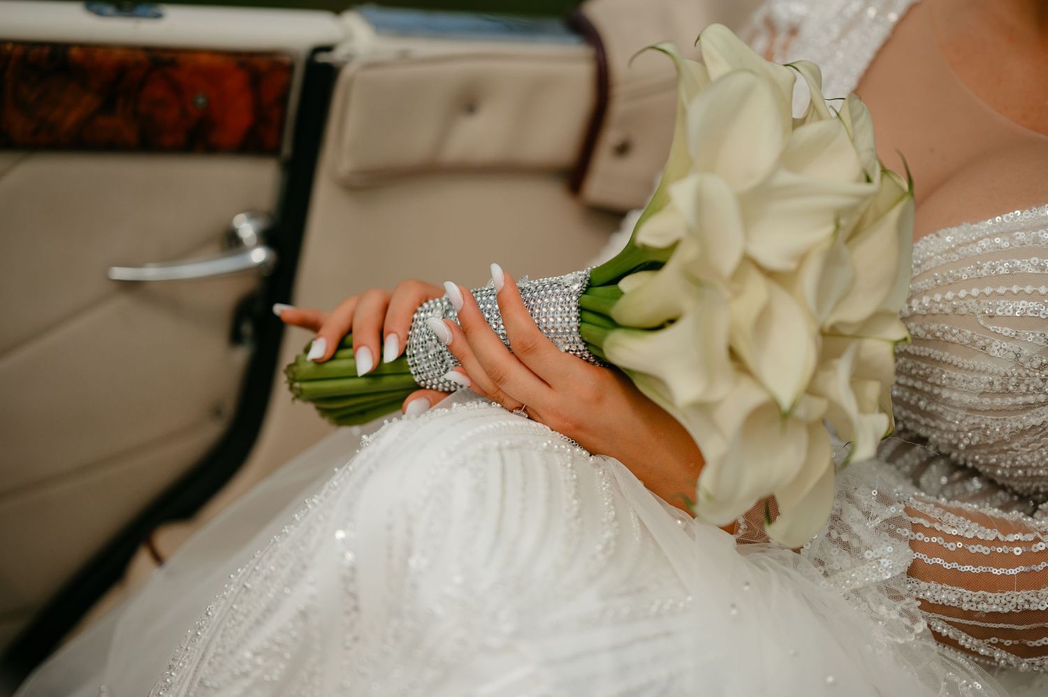 Close-up of bride holding white calla lily bouquet in sparkling beaded wedding dress seated in vintage car.