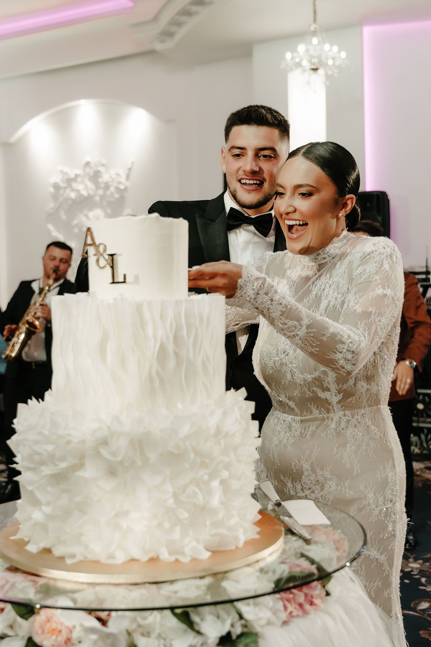 Joyful couple cutting their white feathered wedding cake with gold initials at their reception.