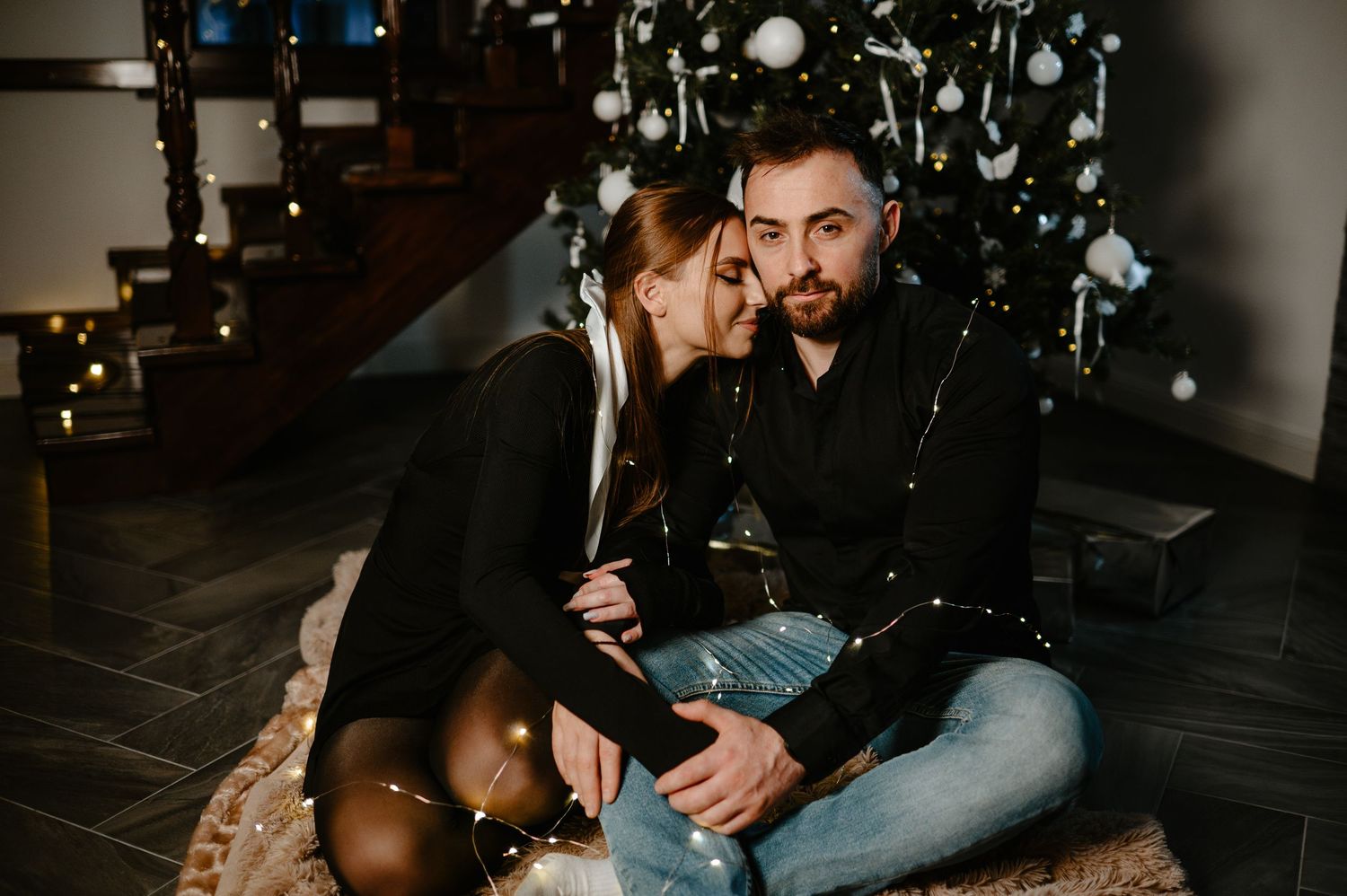 Couple sitting on a fur rug near a Christmas tree with string lights holding a gift.