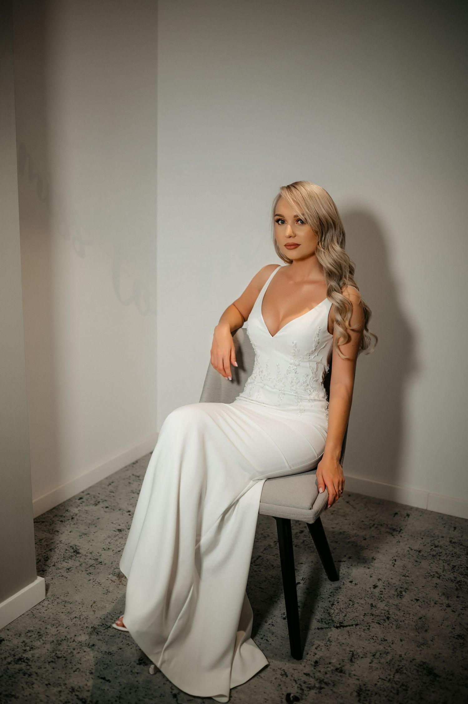 Elegant blonde bride in a white V-neck wedding gown seated gracefully on a chair in a bright room.