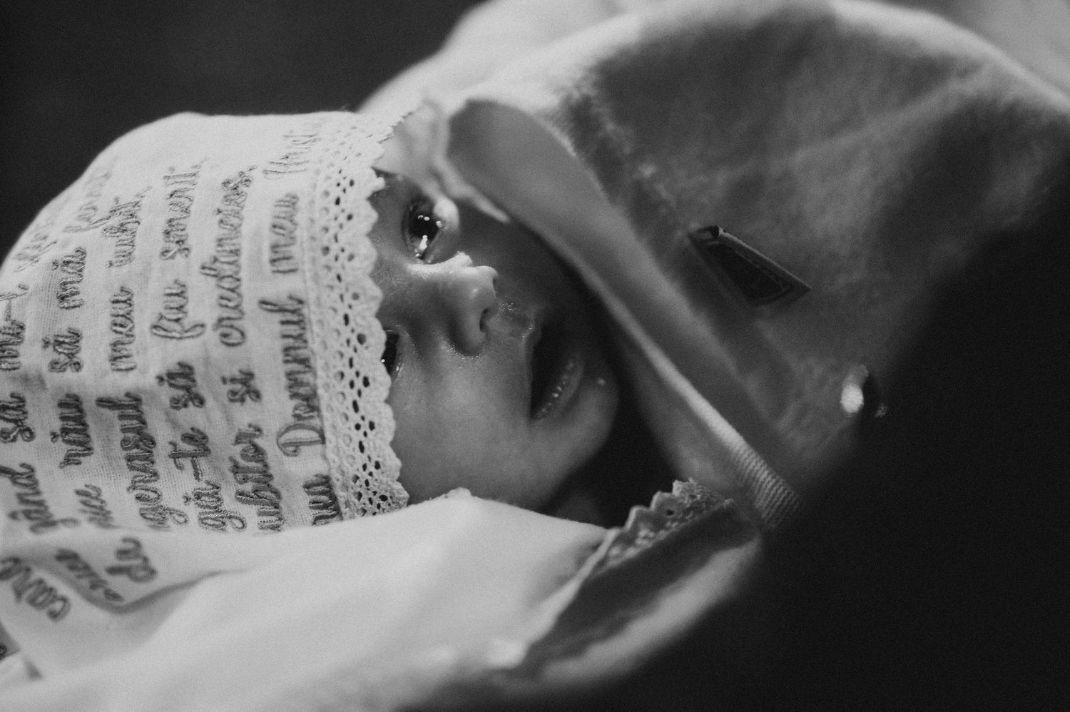 Black and white close-up of newborn in baptism bonnet with religious text embroidery.