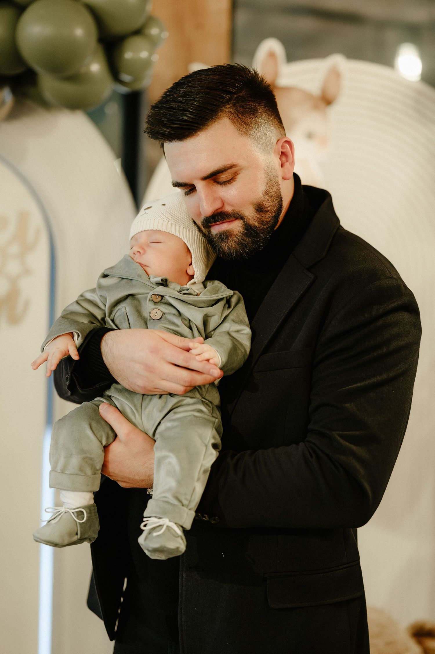 Loving father cradling his sleeping newborn baby dressed in a sage green outfit at a christening party.