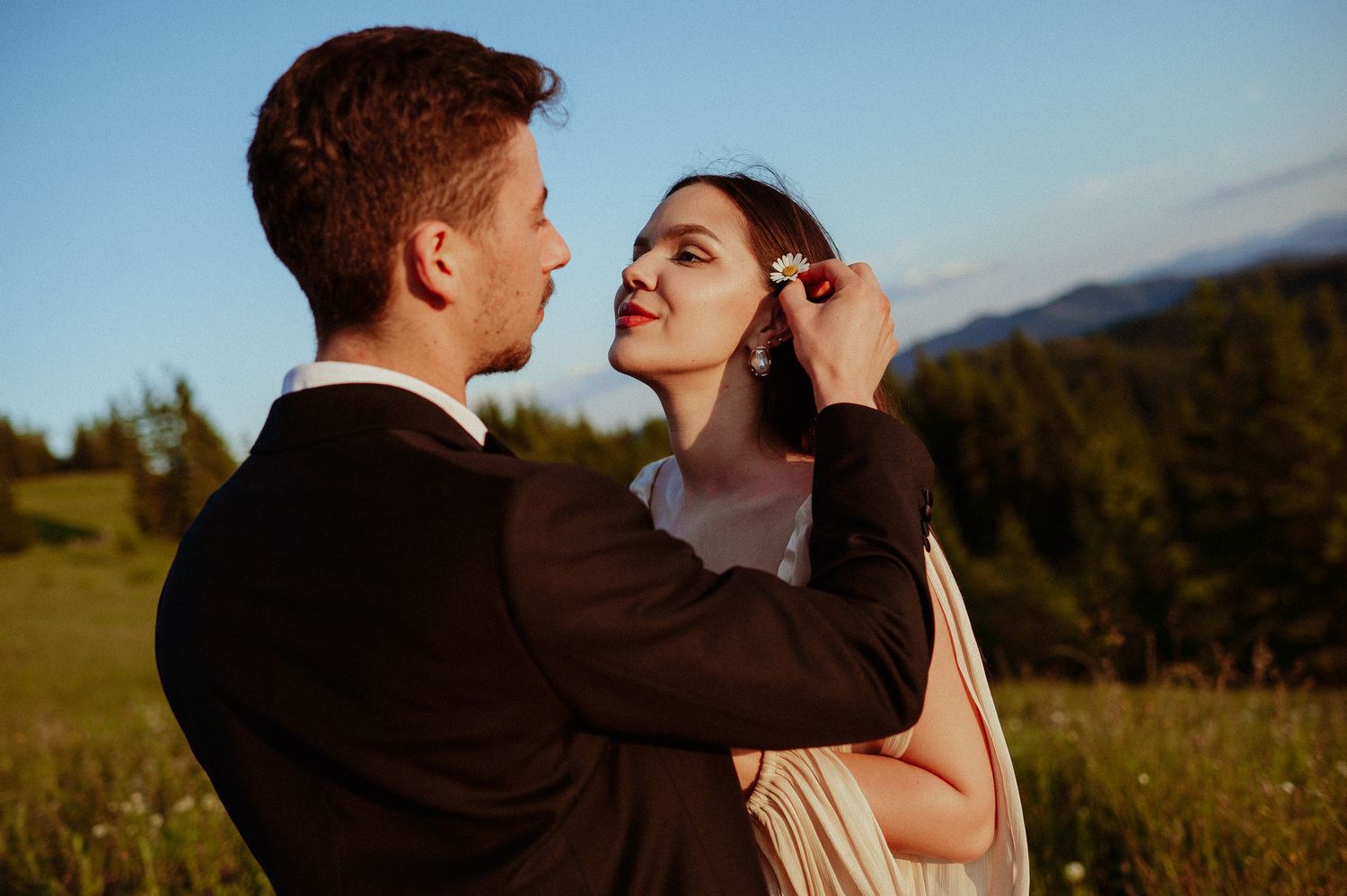 Groom tenderly tucking hair behind the bride's ear in a golden sunset meadow.