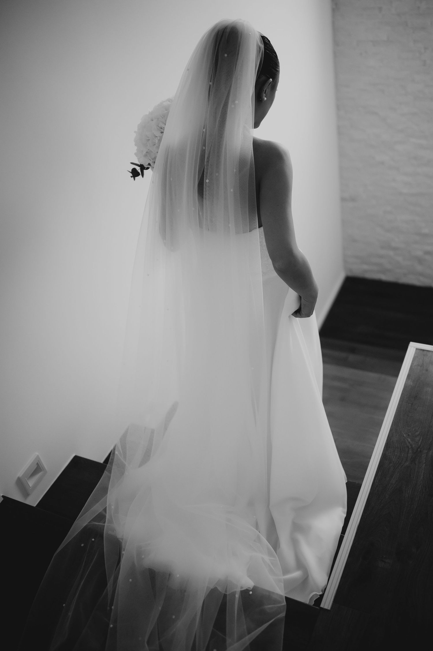 Black and white rear view of bride in flowing wedding gown and long veil on staircase.