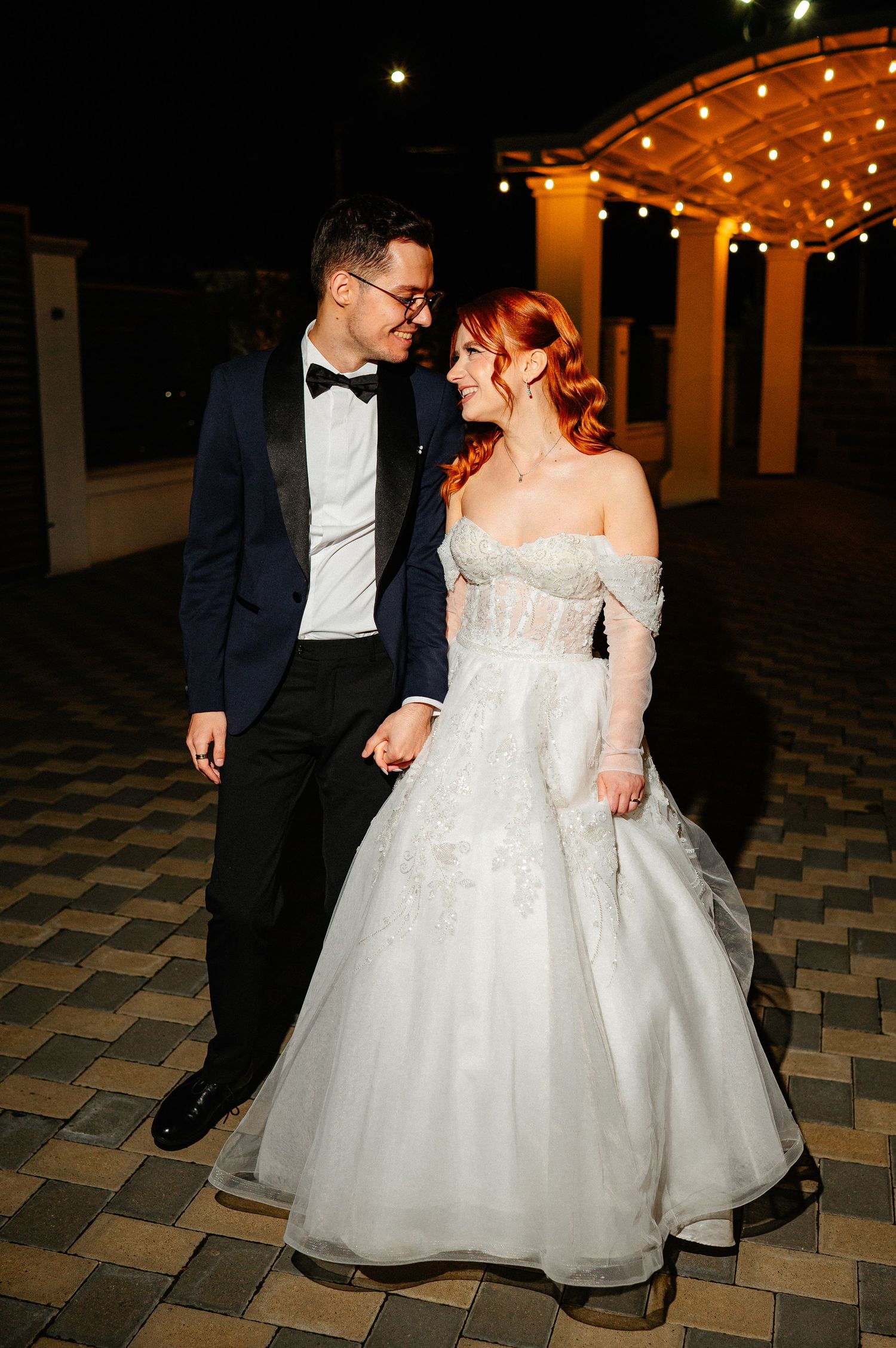 Bride and groom share a tender moment outdoors at night under string lights.