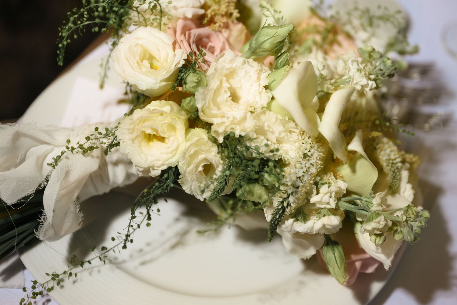 Elegant wedding bouquet featuring cream roses, calla lilies, and delicate greenery arranged on a white surface.