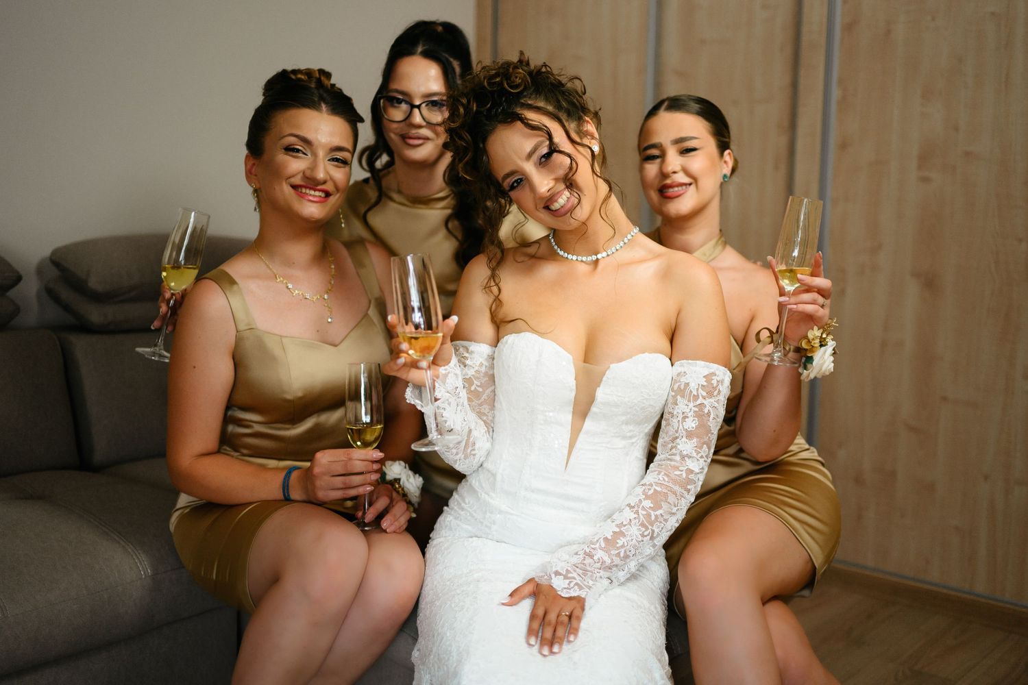 Bride in white lace gown poses with three bridesmaids in gold dresses, holding champagne glasses.