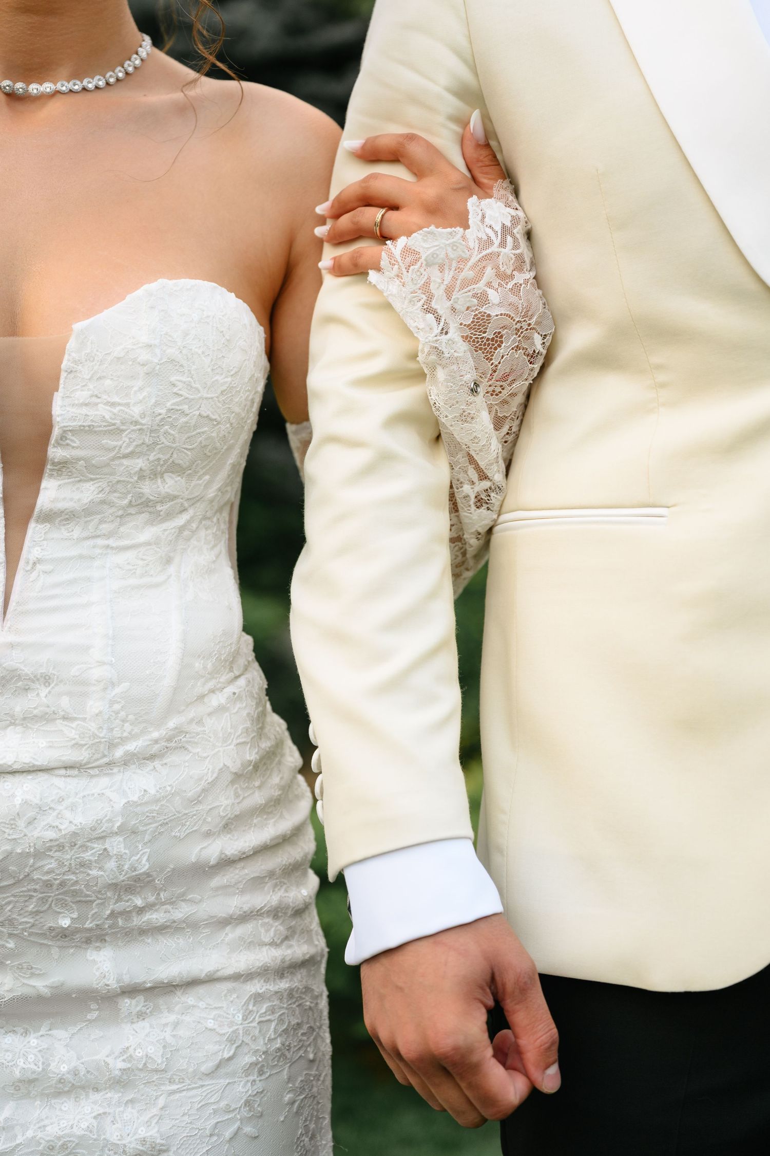 Bride in white lace gown holding groom's arm in cream tuxedo jacket at wedding ceremony.