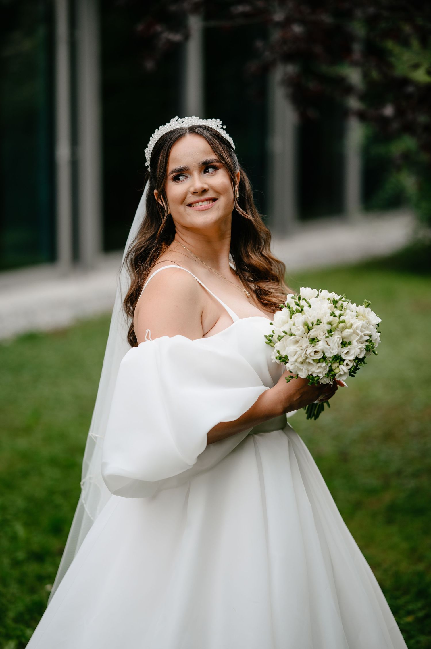 Smiling bride in white off-shoulder gown holding white flower bouquet outdoors.