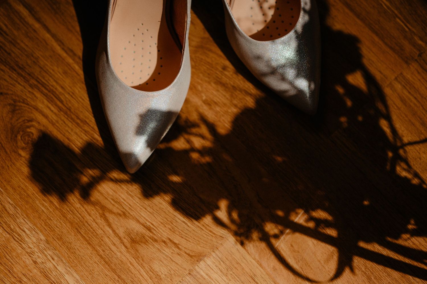 Silver bridal heels casting elegant floral shadows on warm wooden floor in golden light.