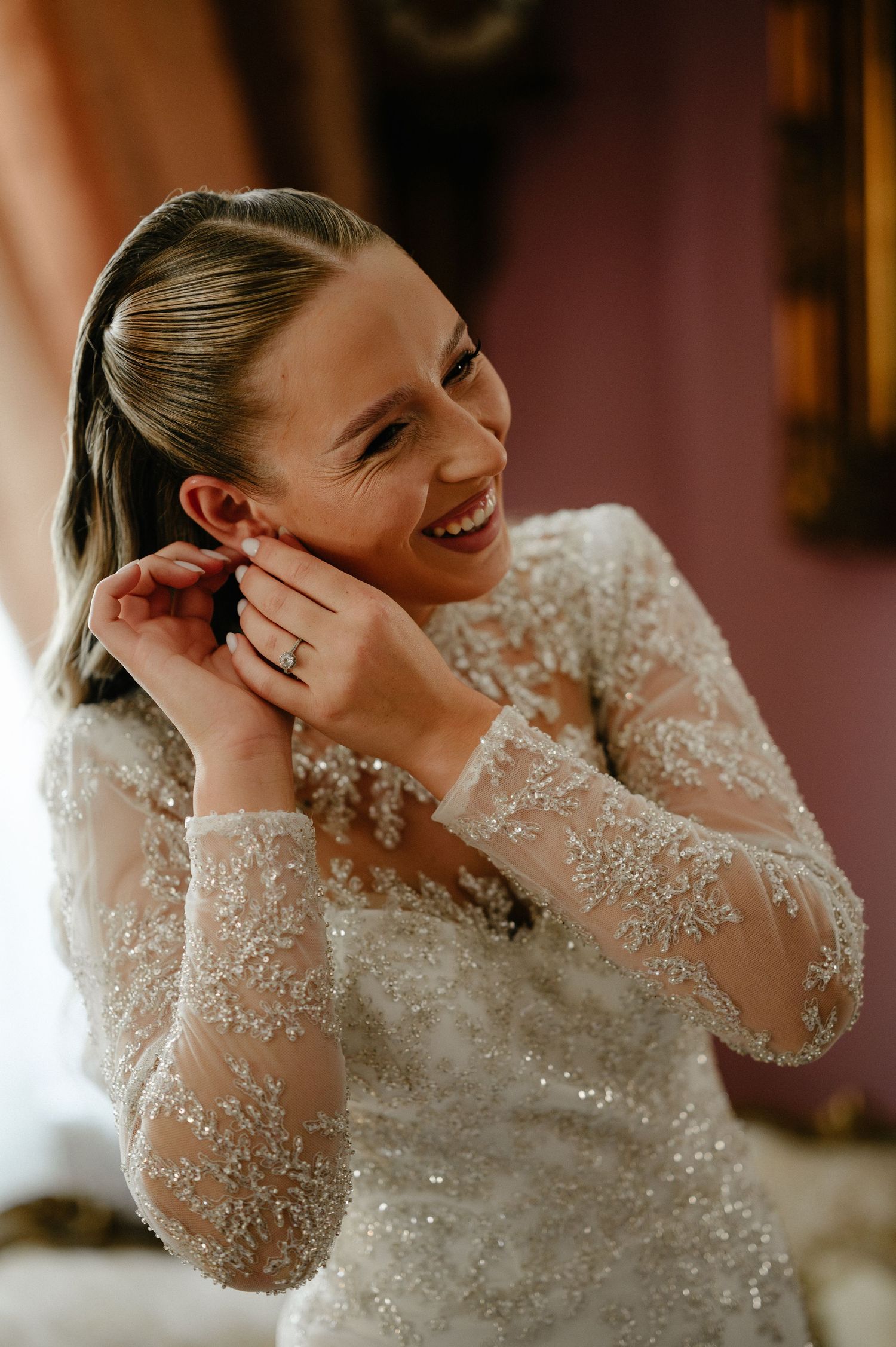 Smiling bride in beaded lace gown adjusting earrings during wedding preparation.