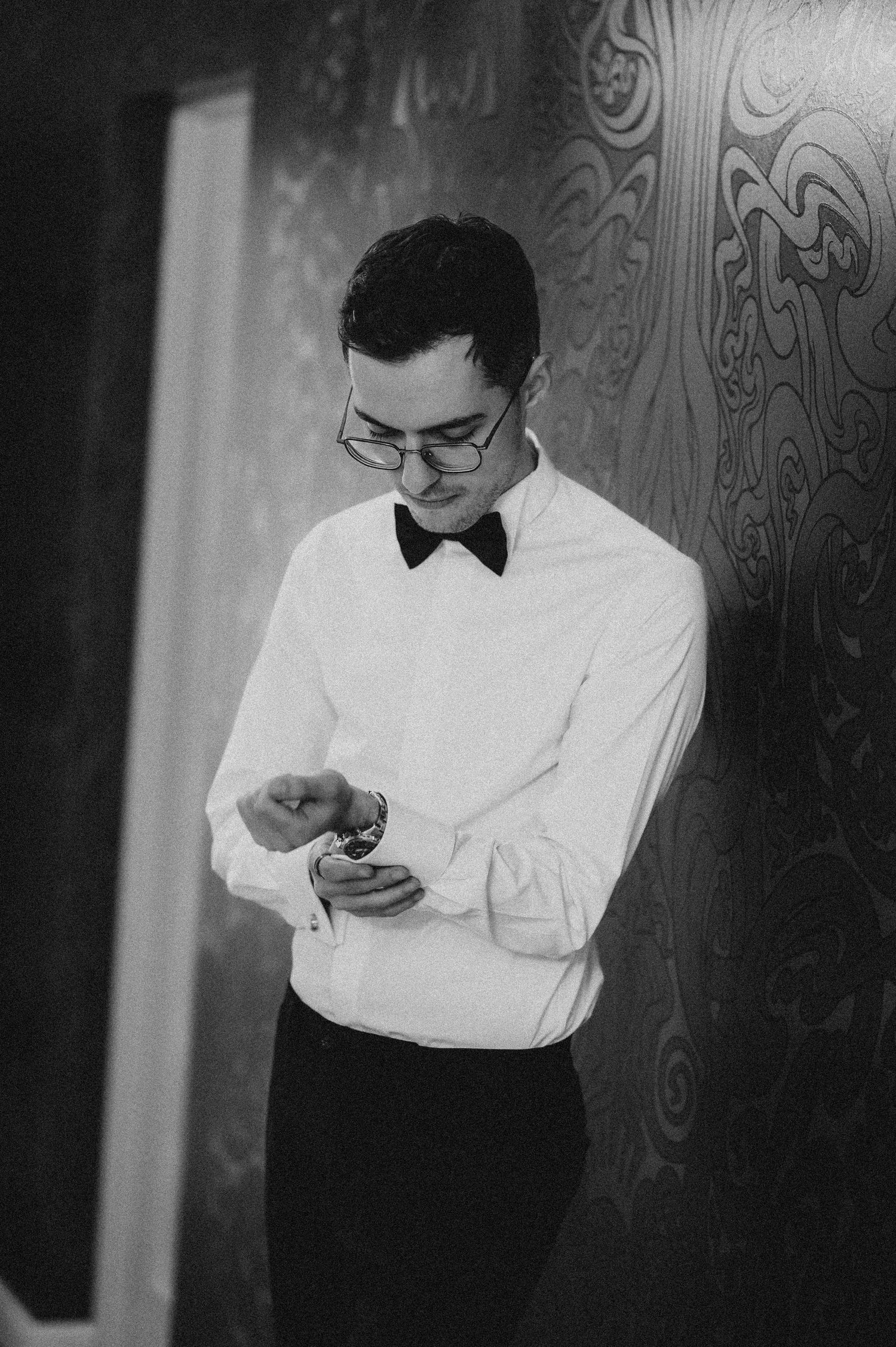 Black and white photo of a young man in a tuxedo shirt and bow tie adjusting his cufflinks.