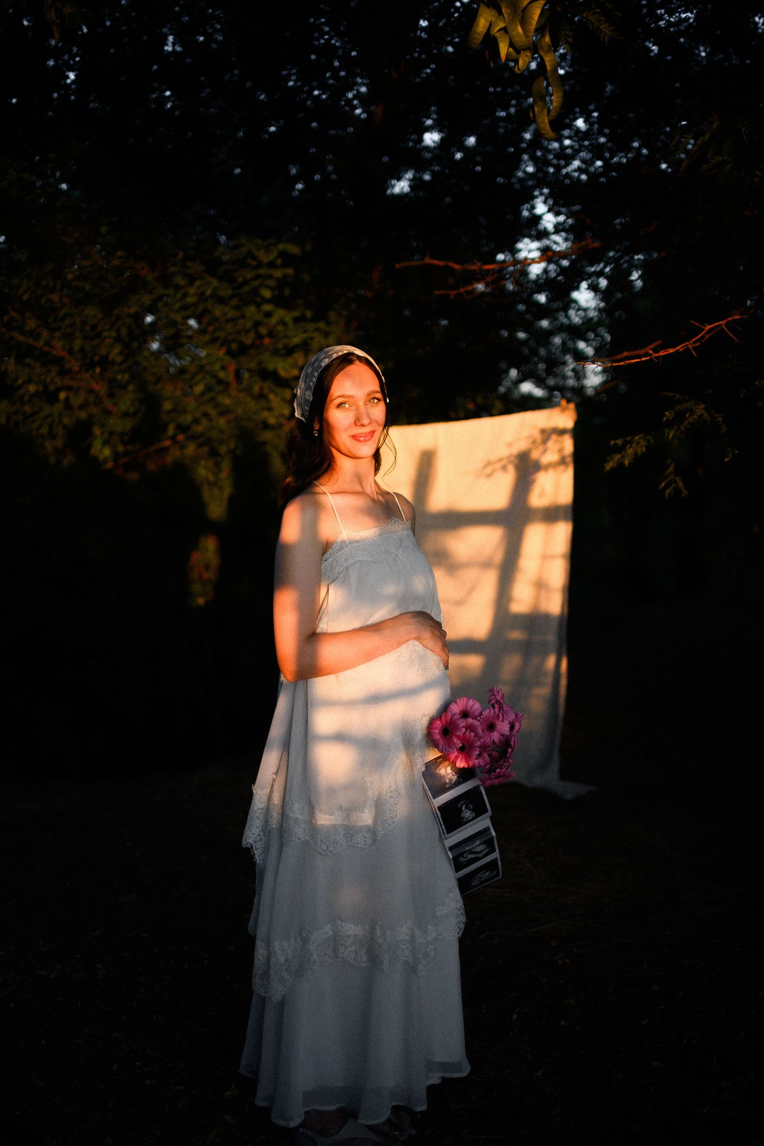 Pregnant woman in white dress holding pink flowers, bathed in golden light outdoors at dusk.