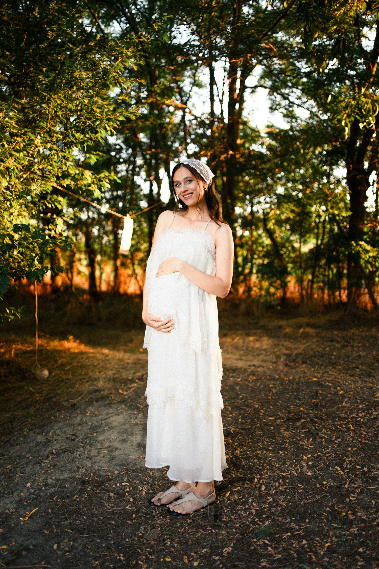Smiling pregnant woman in white dress holding baby bump, standing in sunlit forest during golden hour.