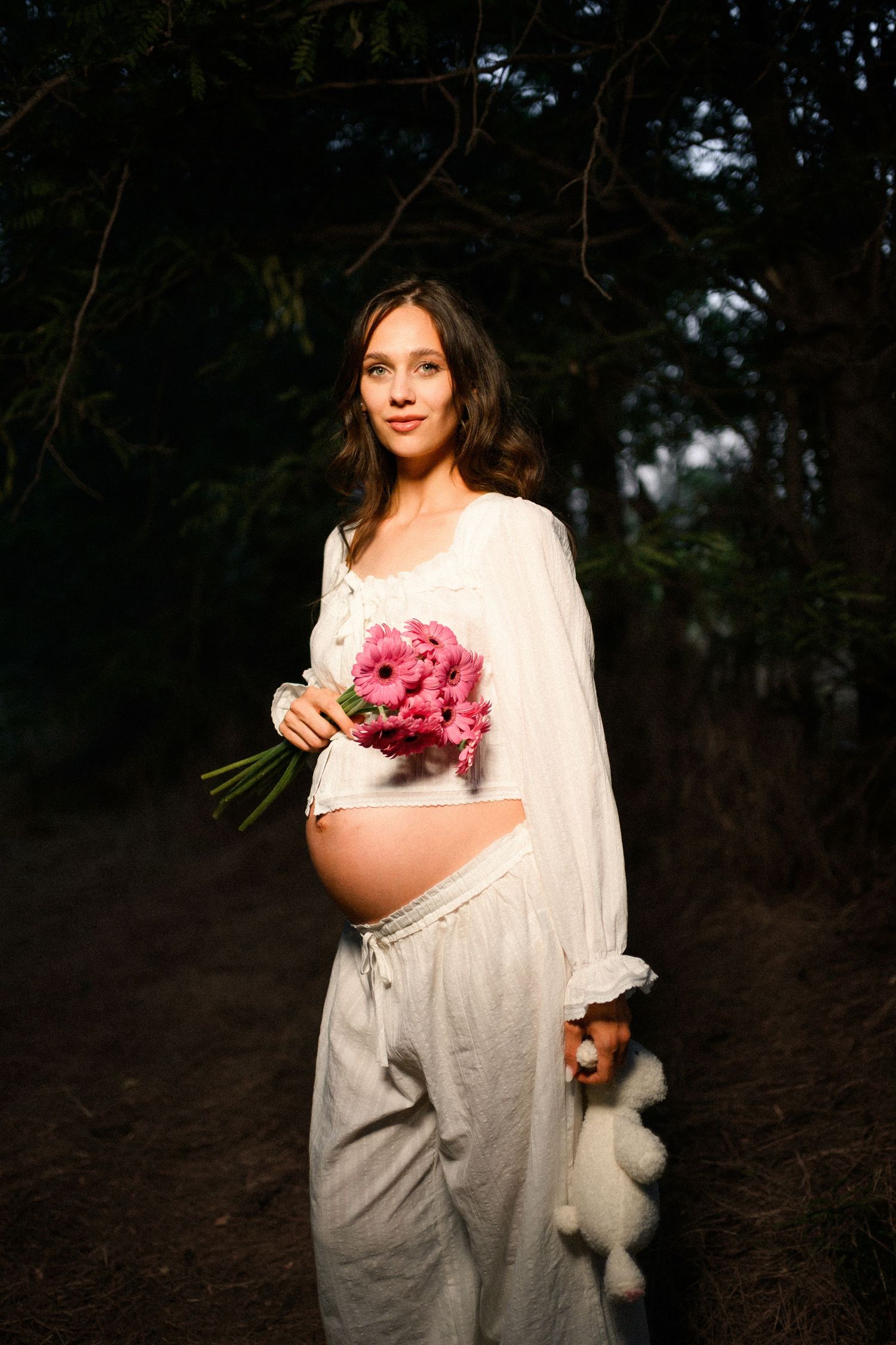 Pregnant woman in white outfit holding pink gerbera bouquet, posing outdoors in a forest setting.