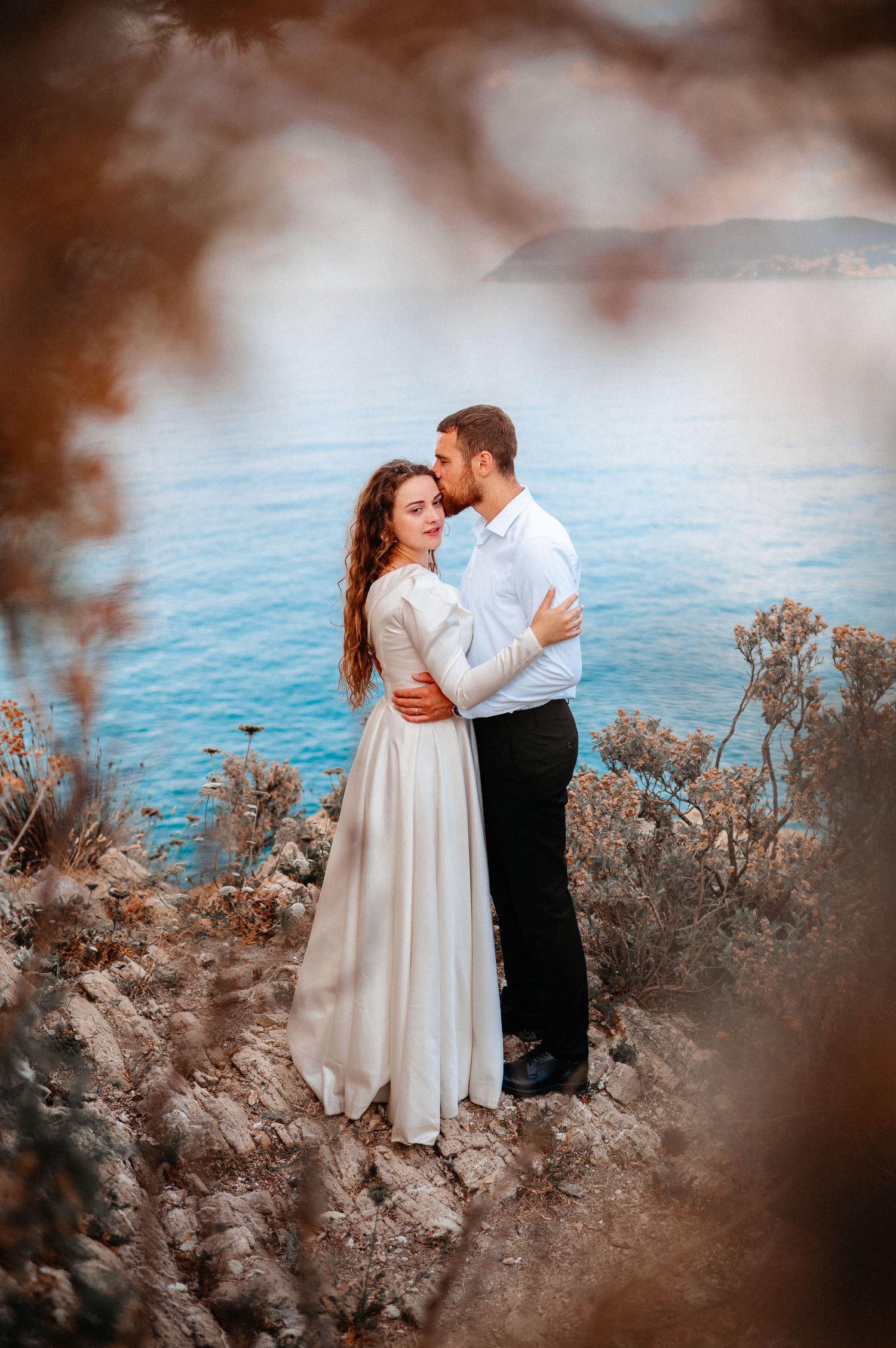 Bride and groom embrace on rocky coastal cliff with turquoise sea backdrop, framed by autumn branches.