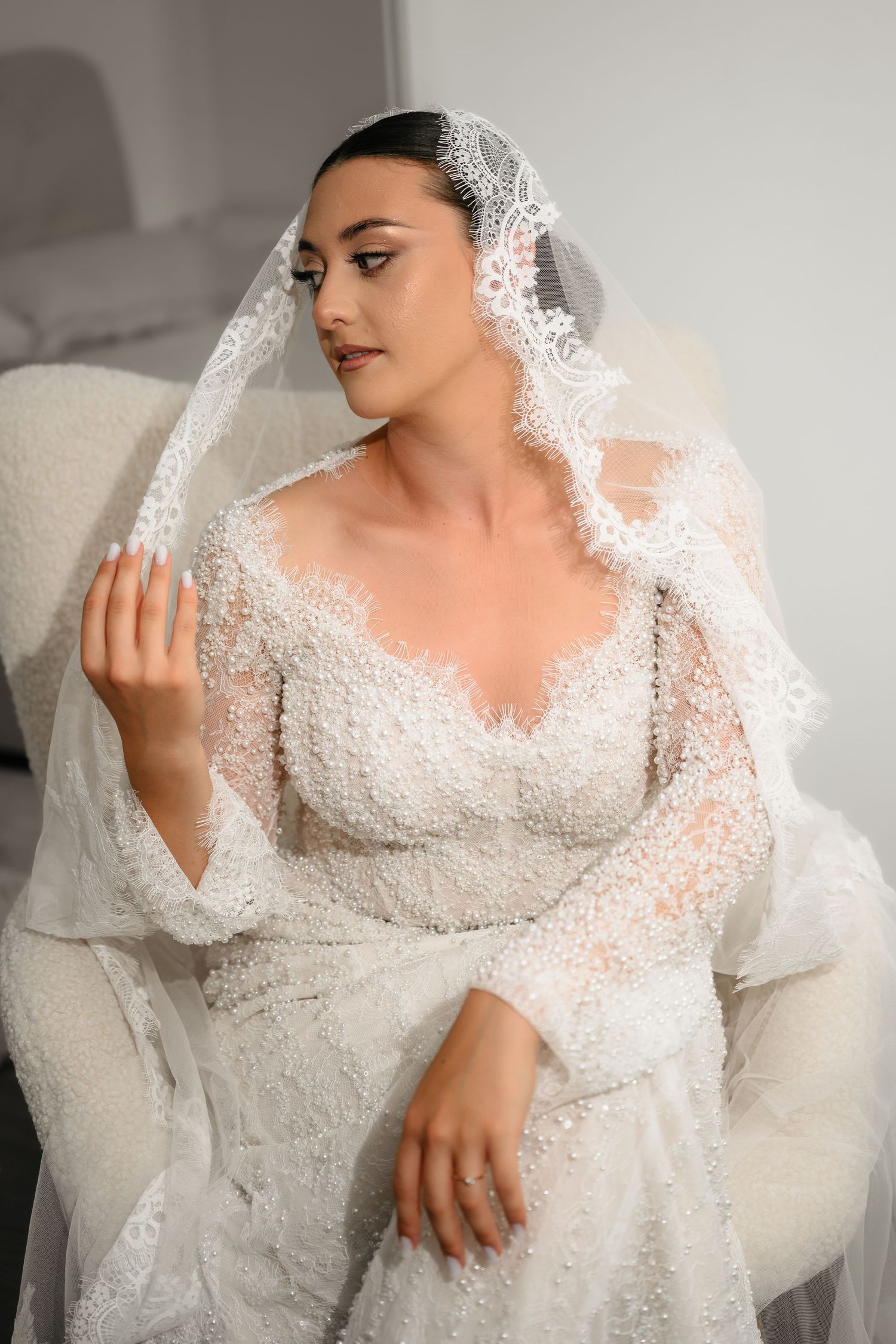 Elegant bride in beaded lace wedding gown and veil seated gracefully in white chair.
