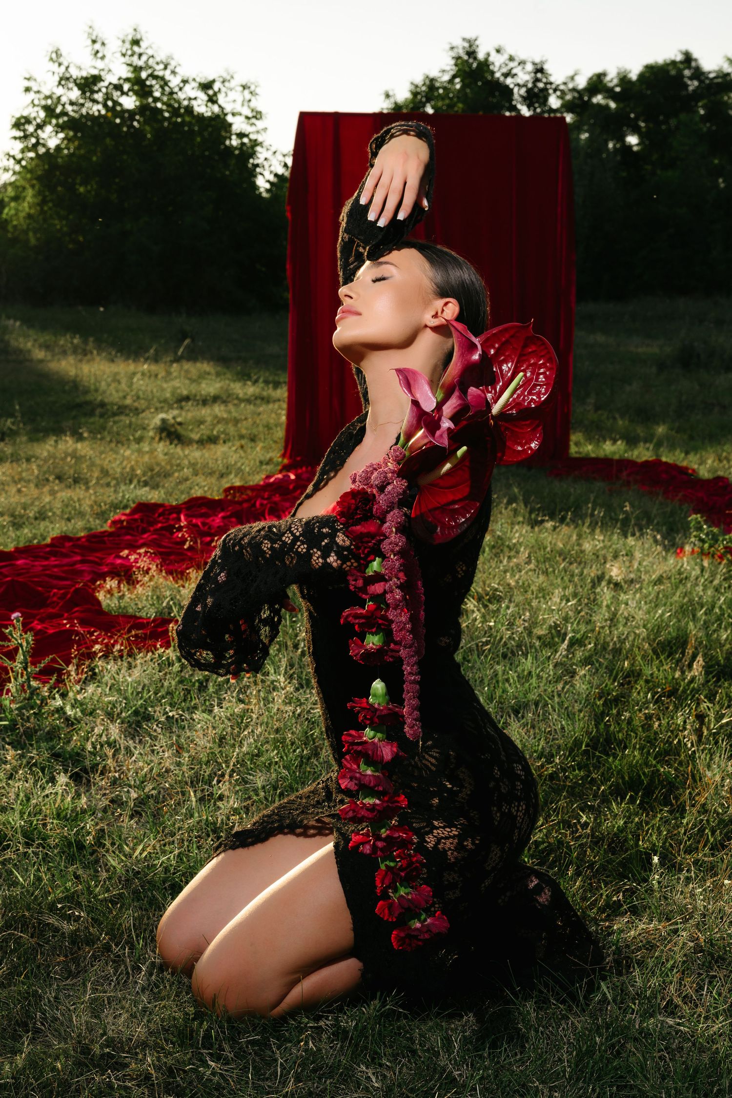 Woman in black lace floral dress kneeling on grass with red fabric backdrop in dramatic outdoor fashion pose.