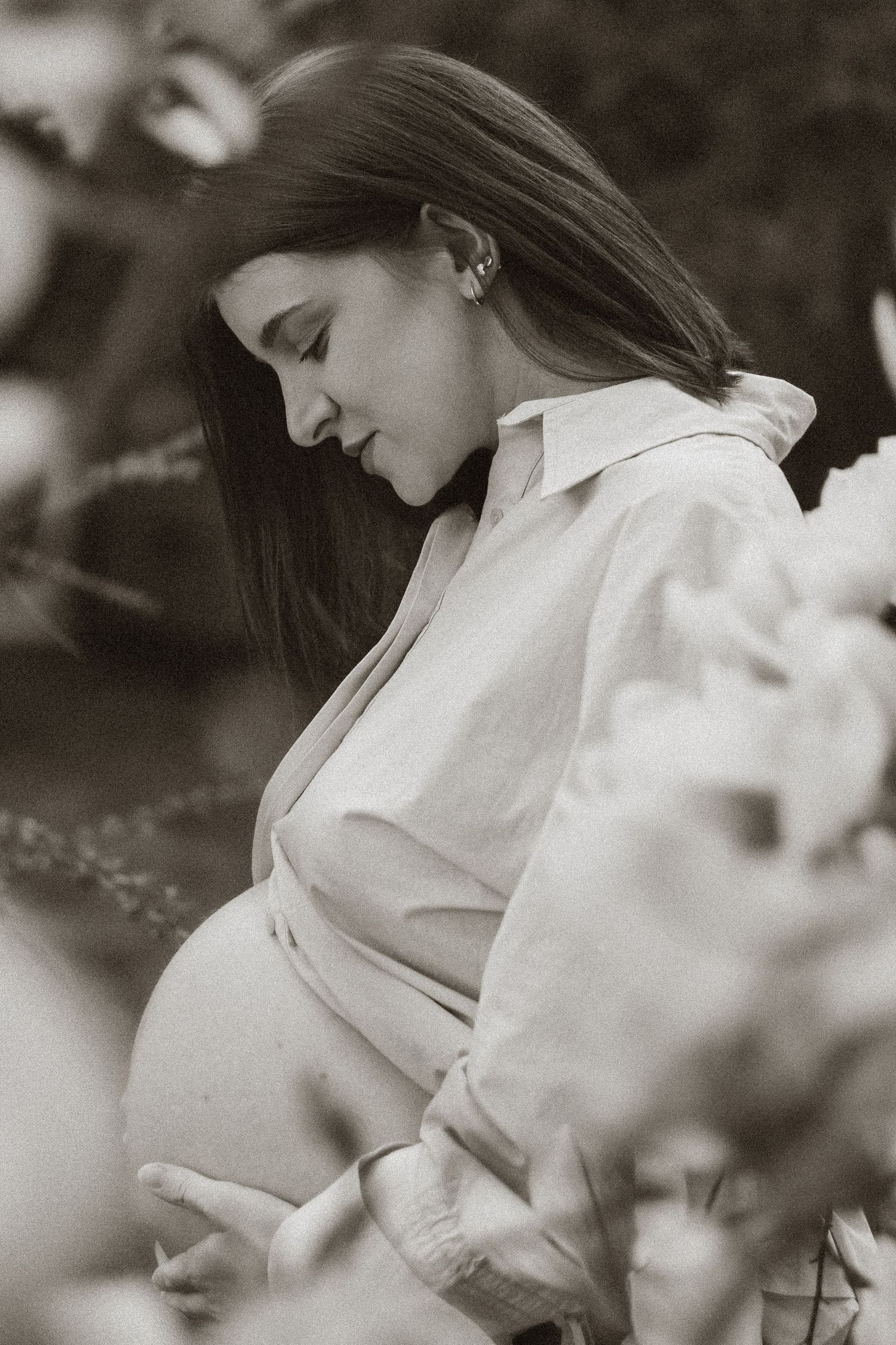 Black and white photo of pregnant woman holding baby bump among flowering branches.