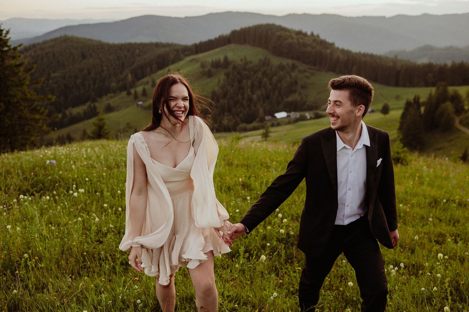 Joyful couple holding hands and laughing while walking through a lush green mountain meadow at sunset.