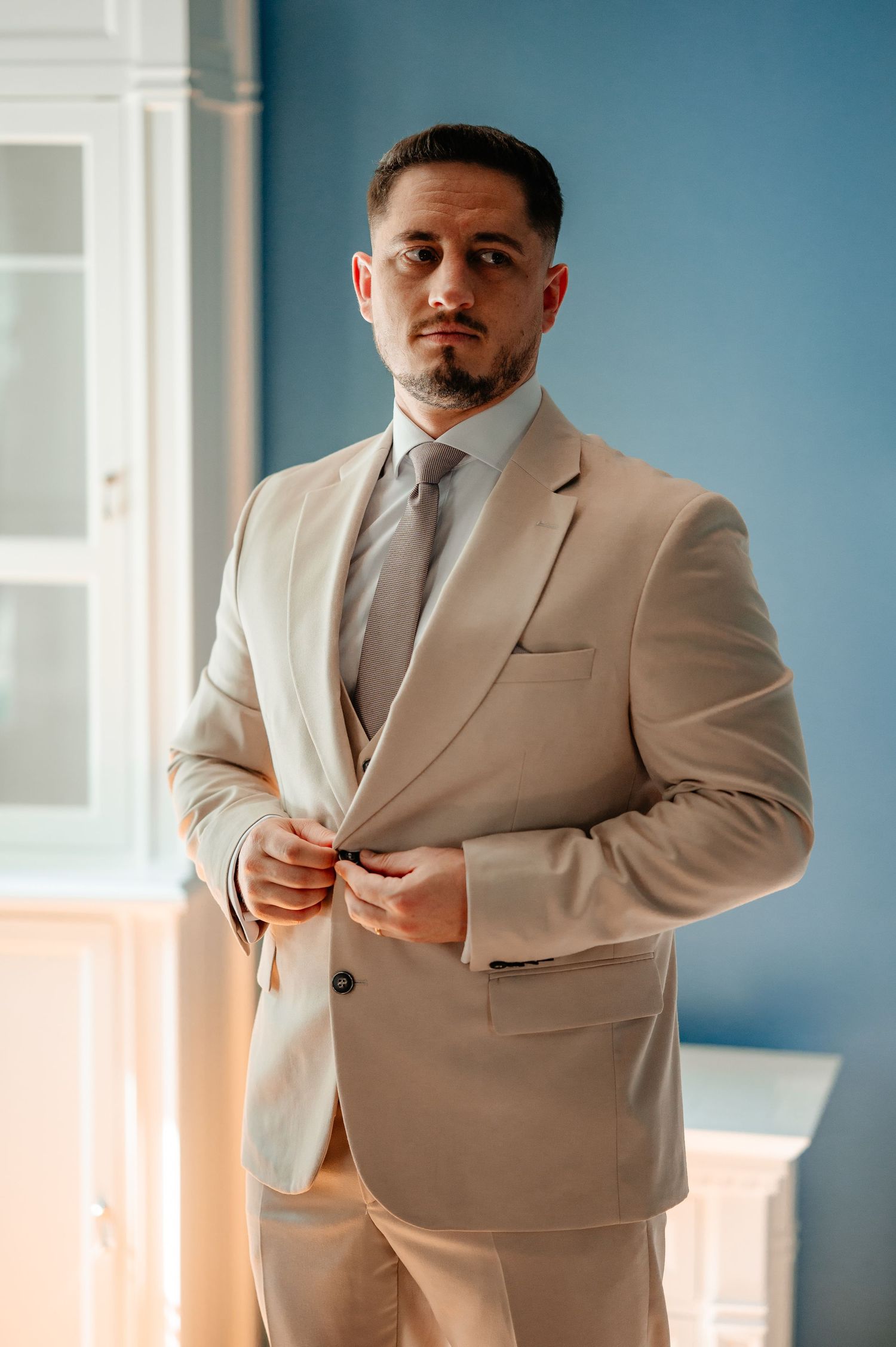 Groom in beige suit buttoning jacket before wedding ceremony.