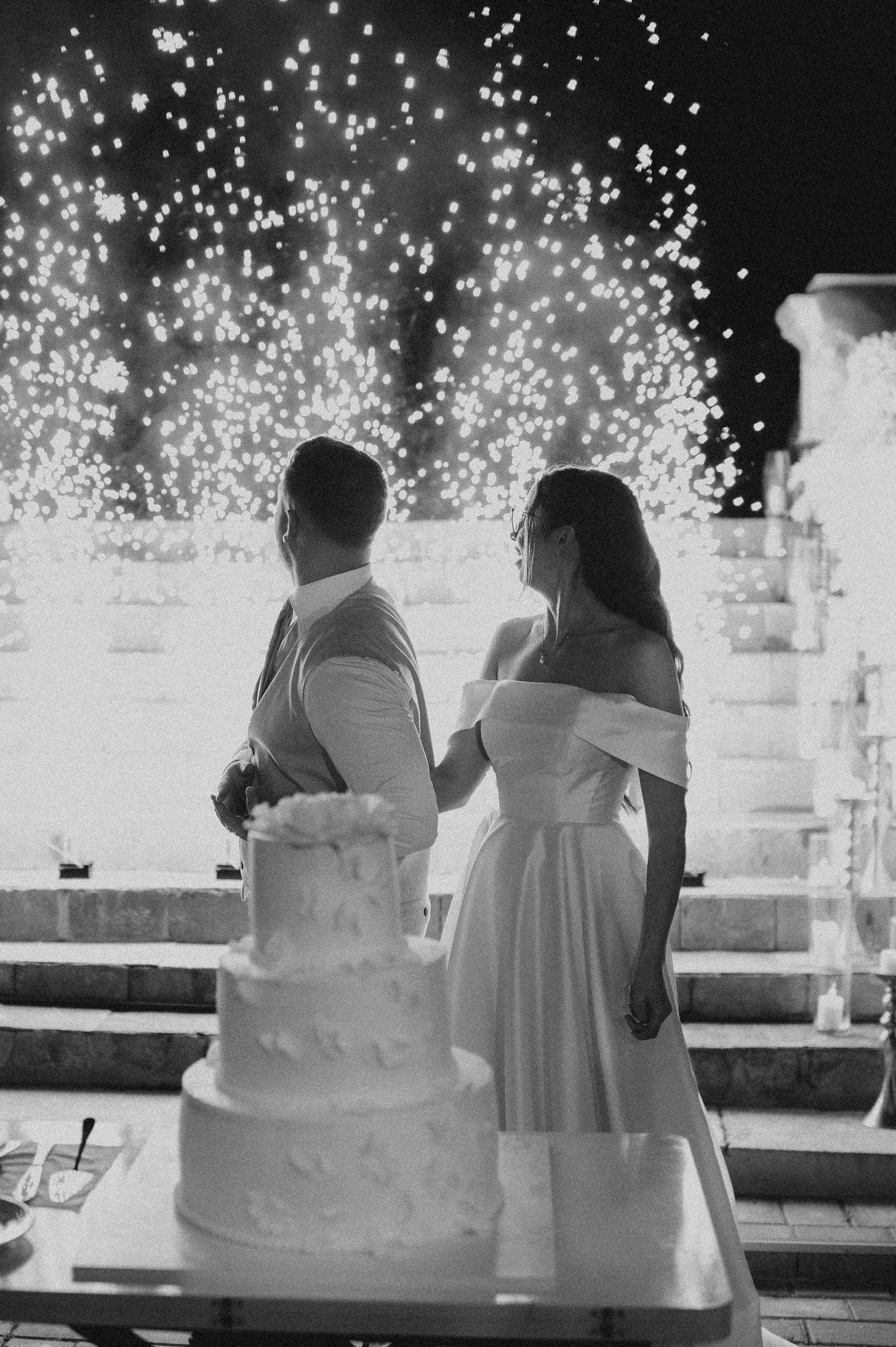 Bride and groom stand before sparkling fireworks display with elegant tiered wedding cake in black and white.