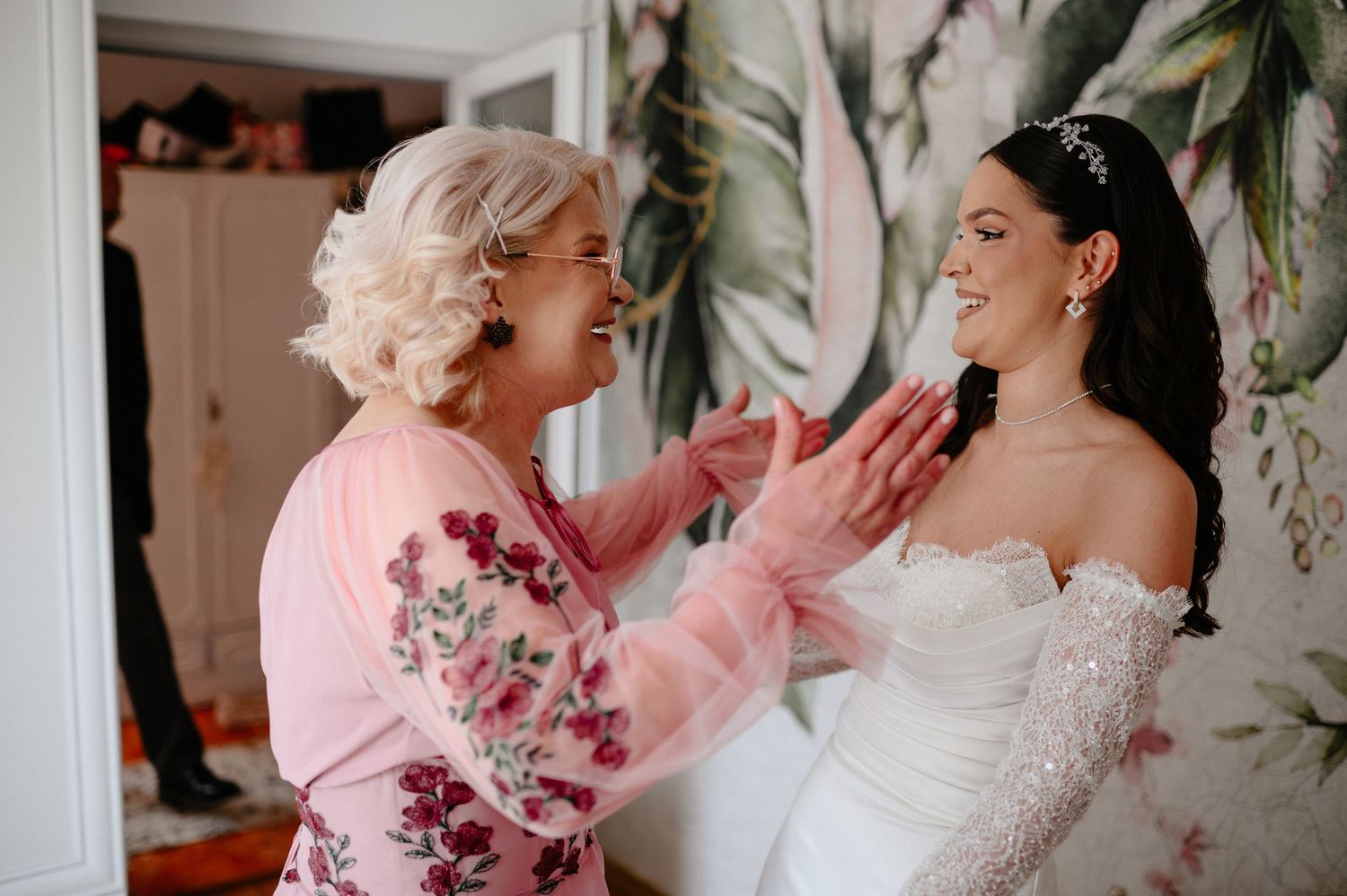 Joyful mother in pink floral dress tenderly touching her smiling daughters face before wedding ceremony.