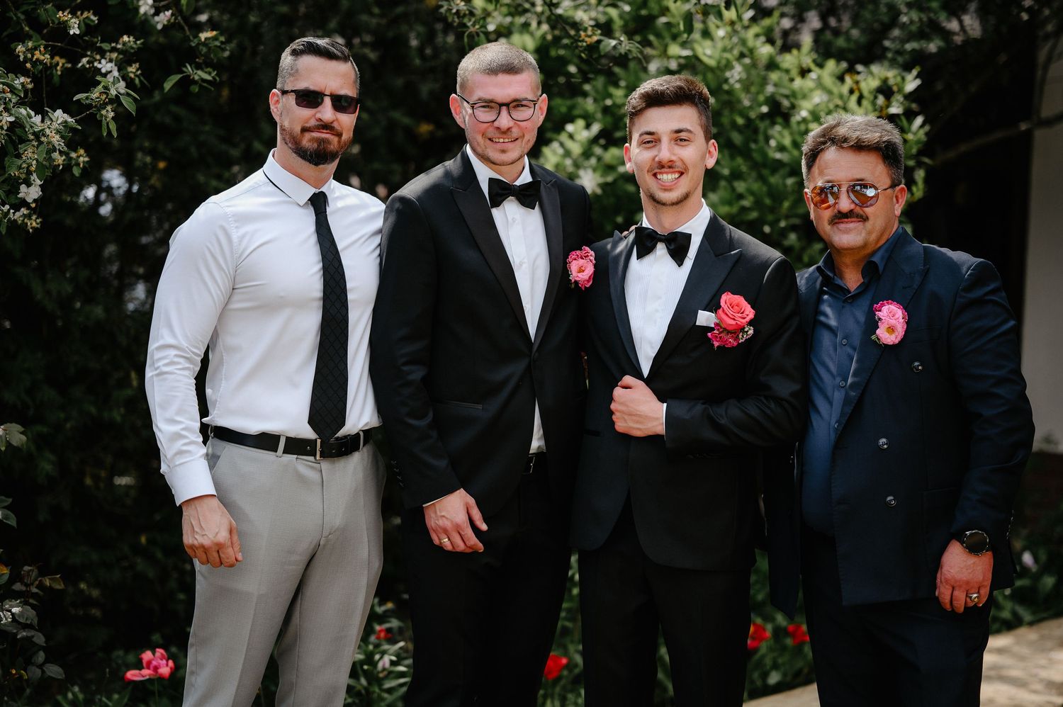 Four groomsmen in formal attire with pink boutonnieres pose together outdoors at a wedding celebration.