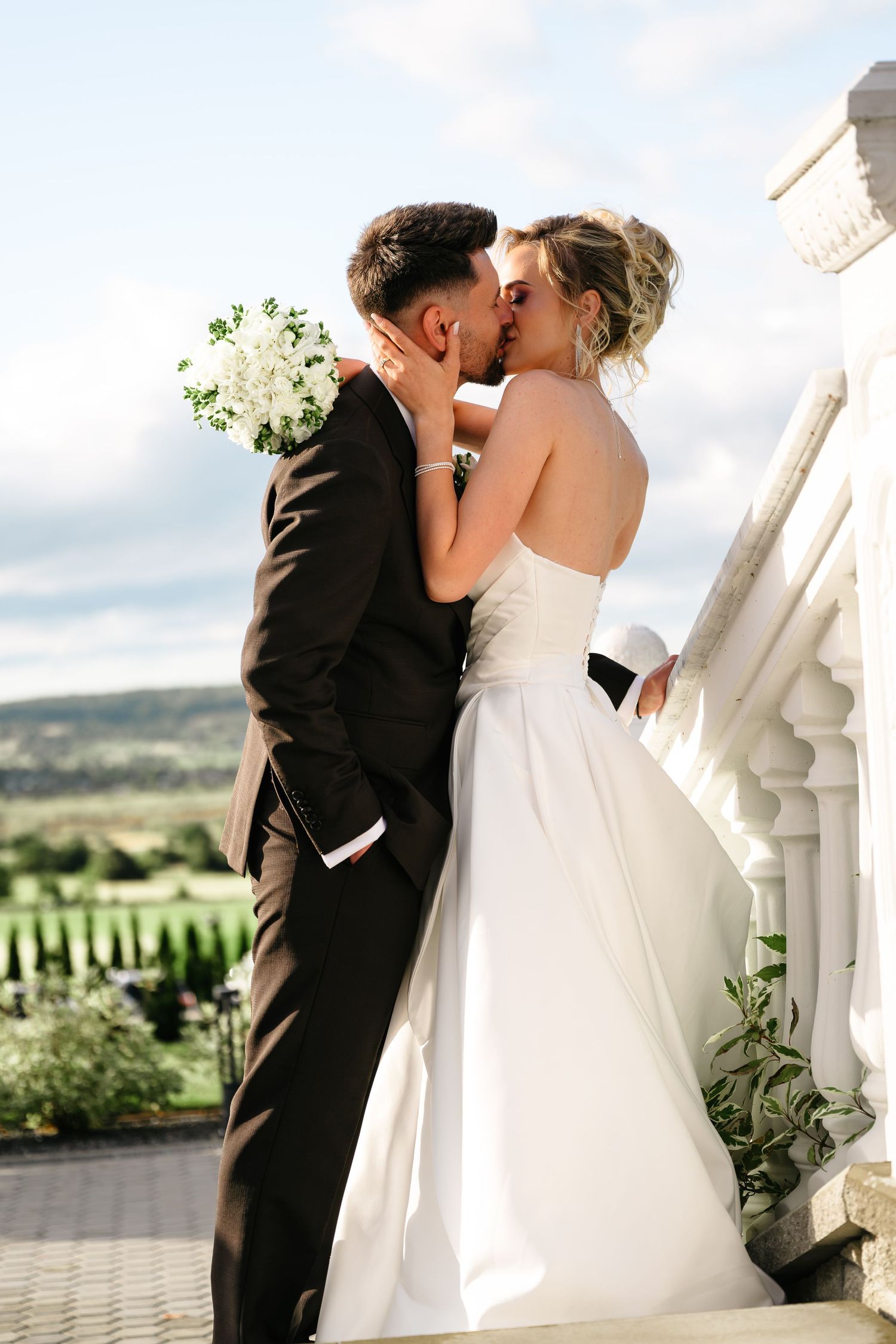 Bride and groom share a romantic kiss on a balcony, with the bride holding a white bouquet on their wedding day.