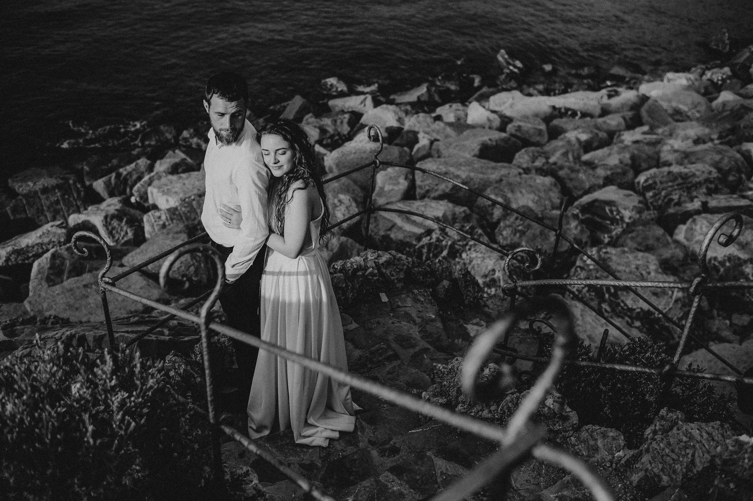 Black and white photo of a couple embracing on rocky seaside steps, smiling together near the ocean.