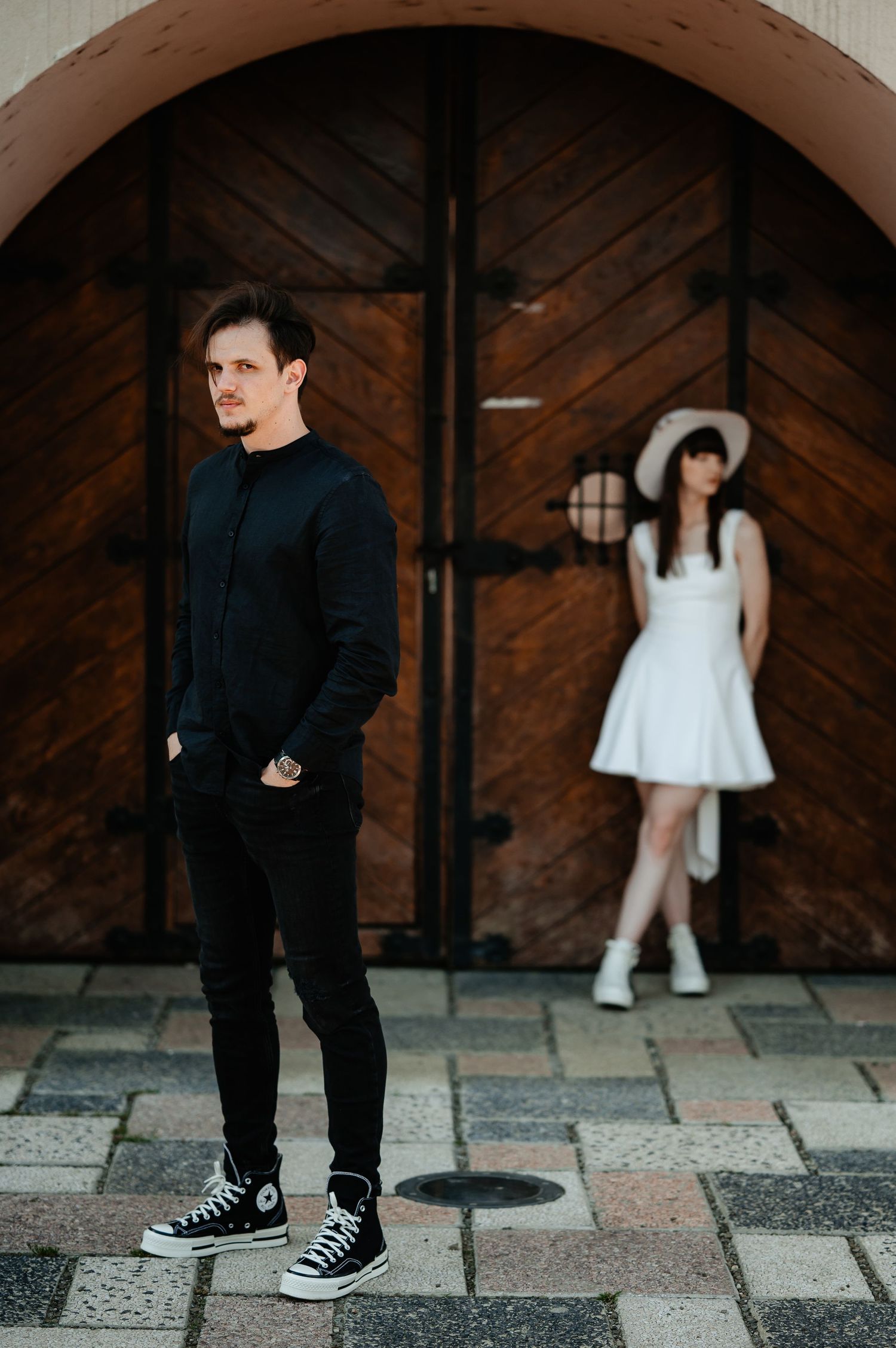 Stylish couple posing apart before large wooden arched doors, man in black, woman in white.