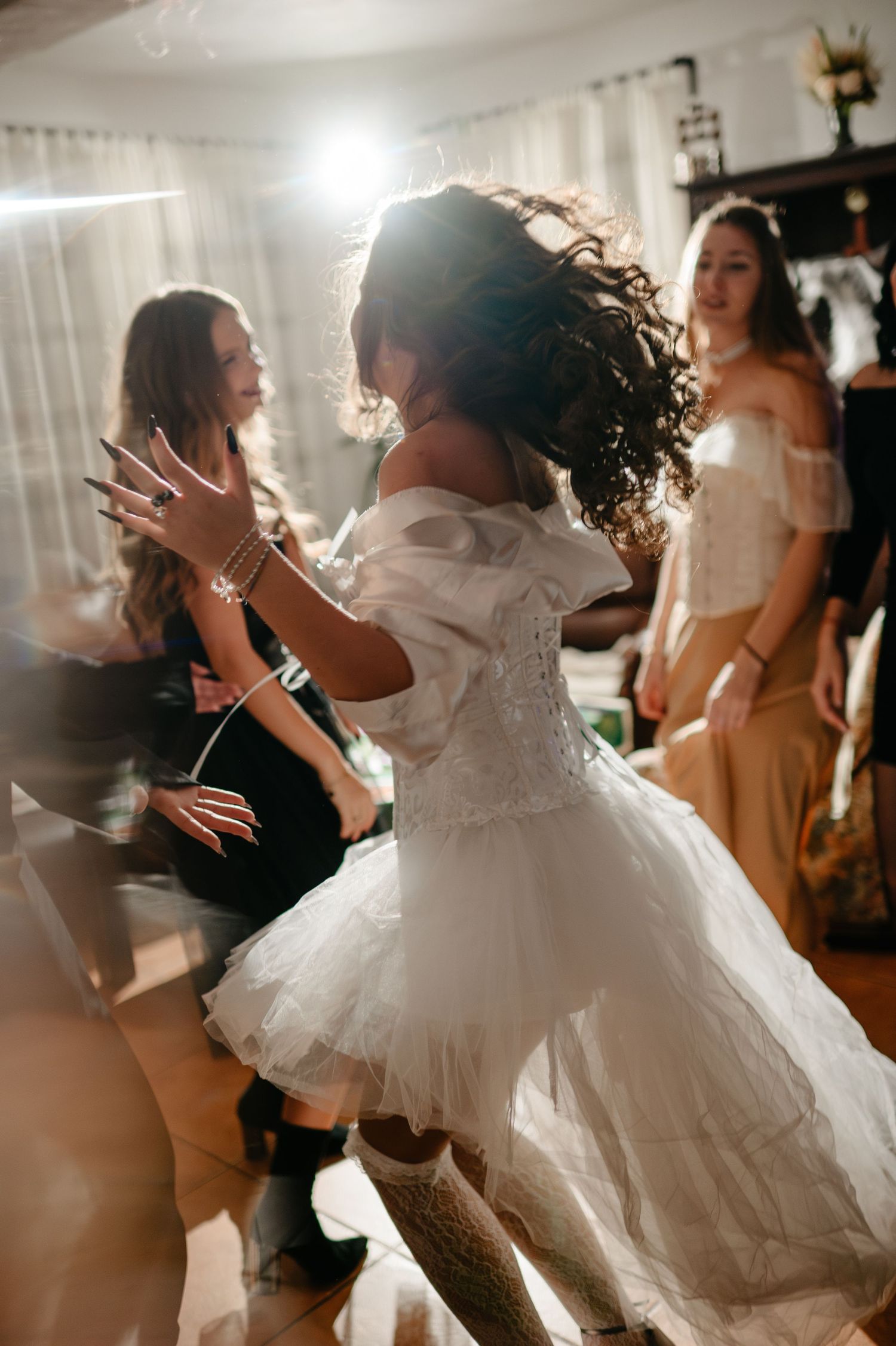 A bride in a white wedding dress twirls and dances joyfully with friends during an indoor celebration.