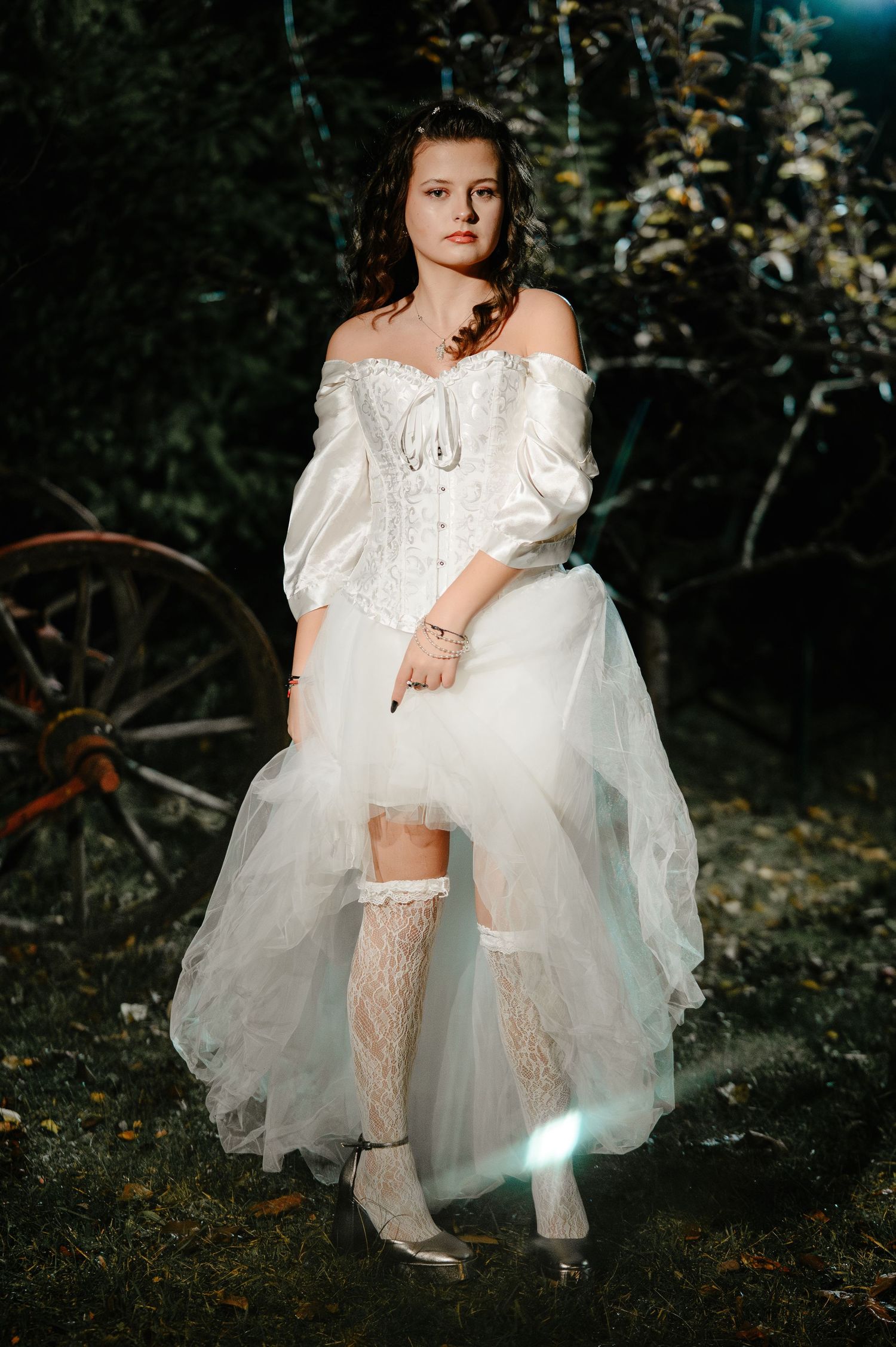 Young woman in white off-shoulder corset gown and lace stockings poses in dark garden setting.