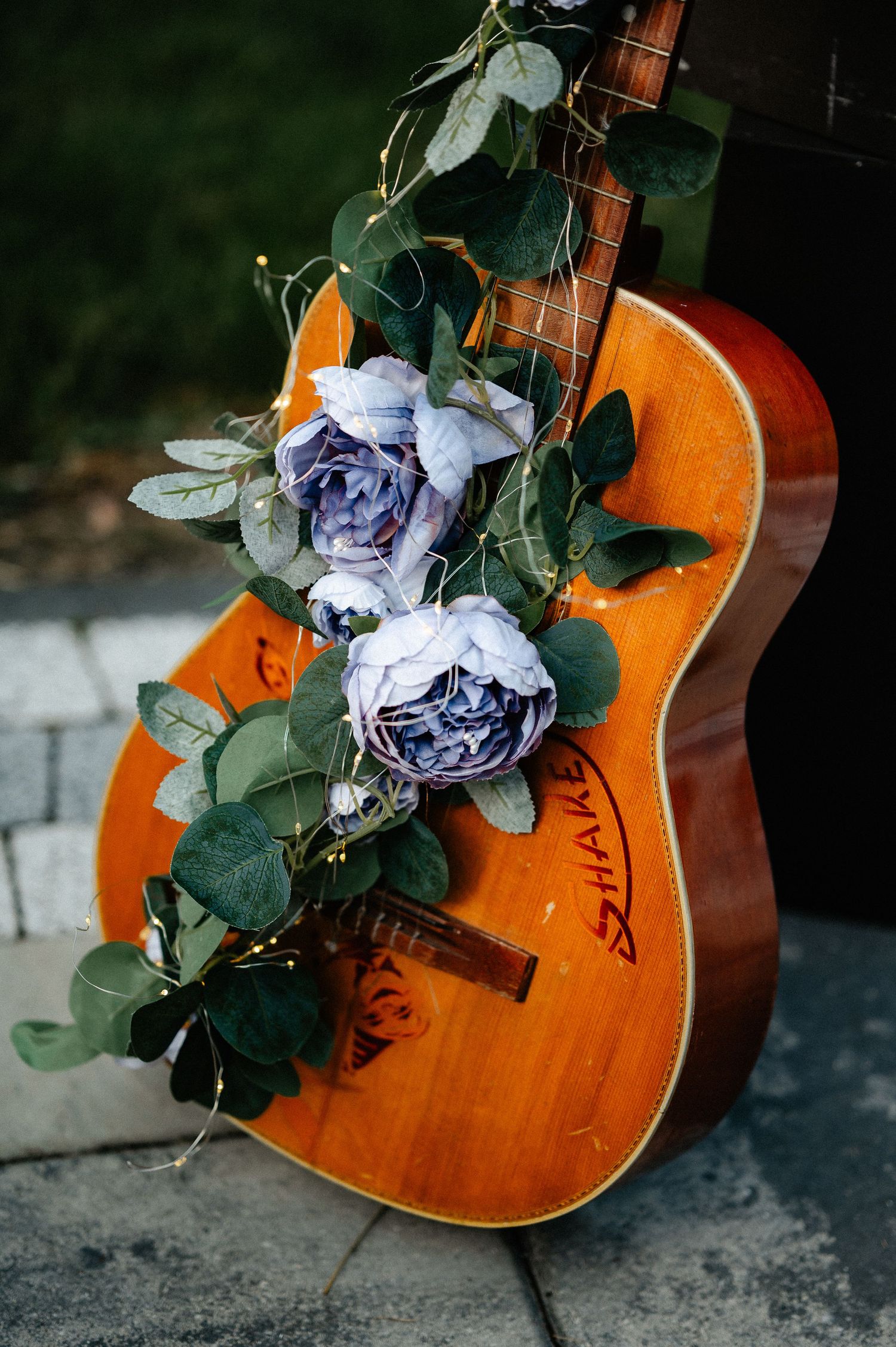 Close-up of acoustic guitar adorned with lavender roses and green eucalyptus leaves with fairy lights.
