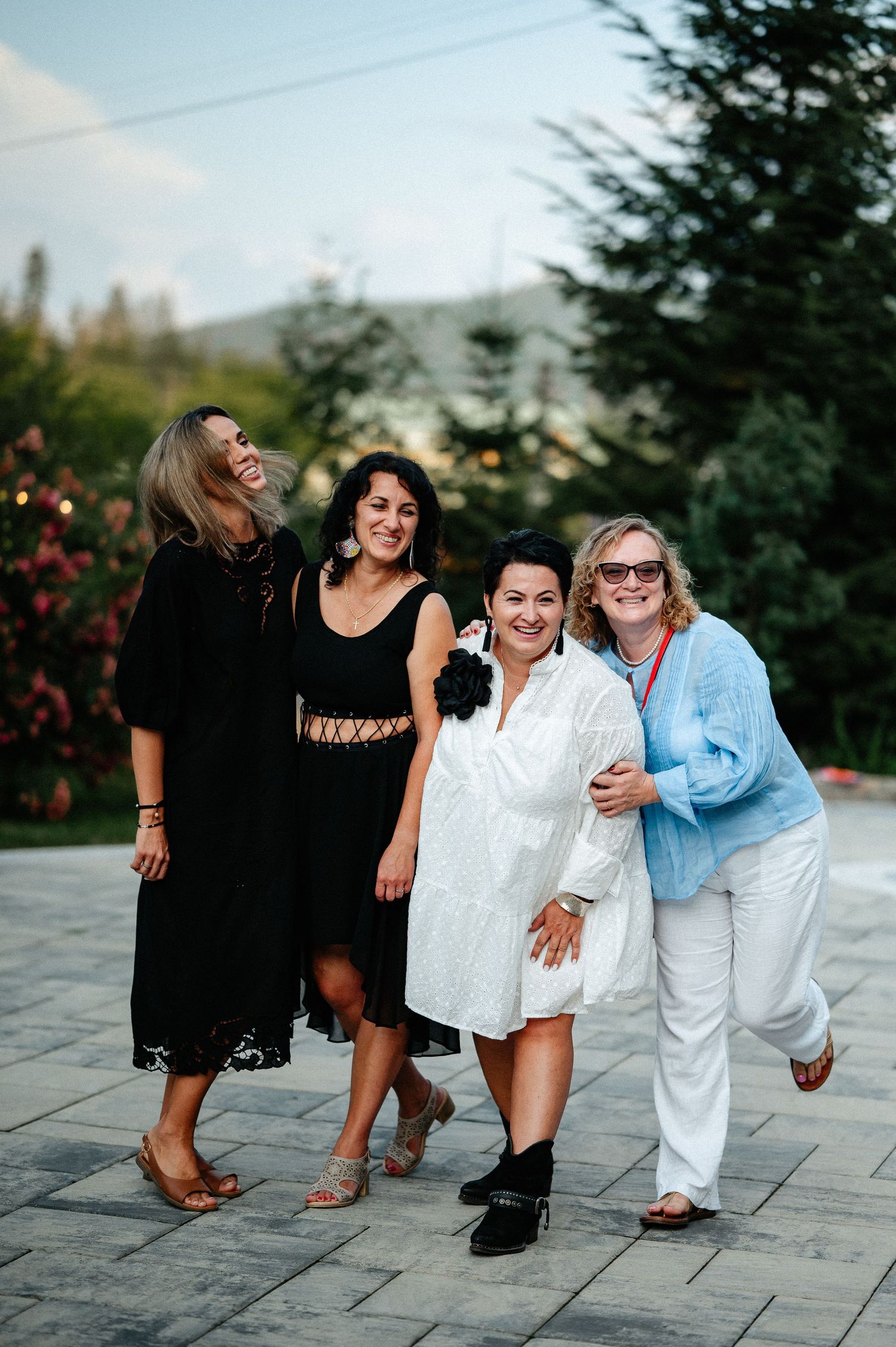 Four women laughing and embracing each other at an outdoor garden event at dusk.